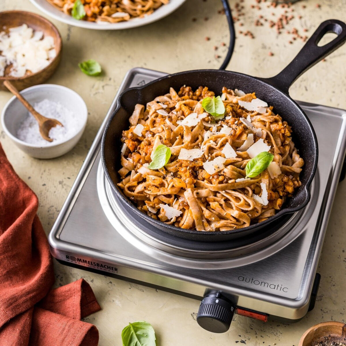 Makaron w czarnej patelni na płycie grzewczej. Ser parmezan, bazylia i sól w pobliżu. Czerwona szmatka po lewej.