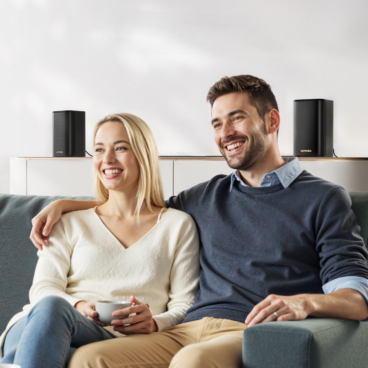 Para uśmiecha się na kanapie. Dwa głośniki na półce. Kobieta z kubkiem. Mężczyzna ją obejmuje.