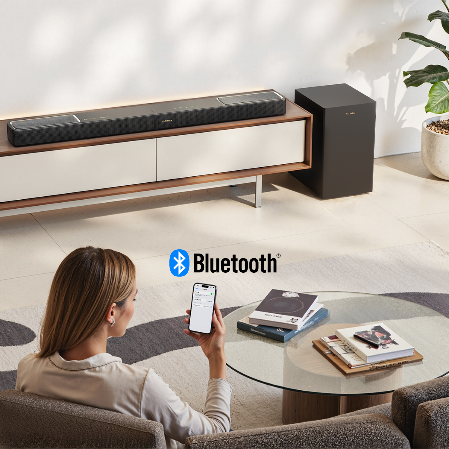 Kobieta łączy się z soundbarem przez Bluetooth w telefonie. Soundbar i subwoofer na szafce. Książki na stole.