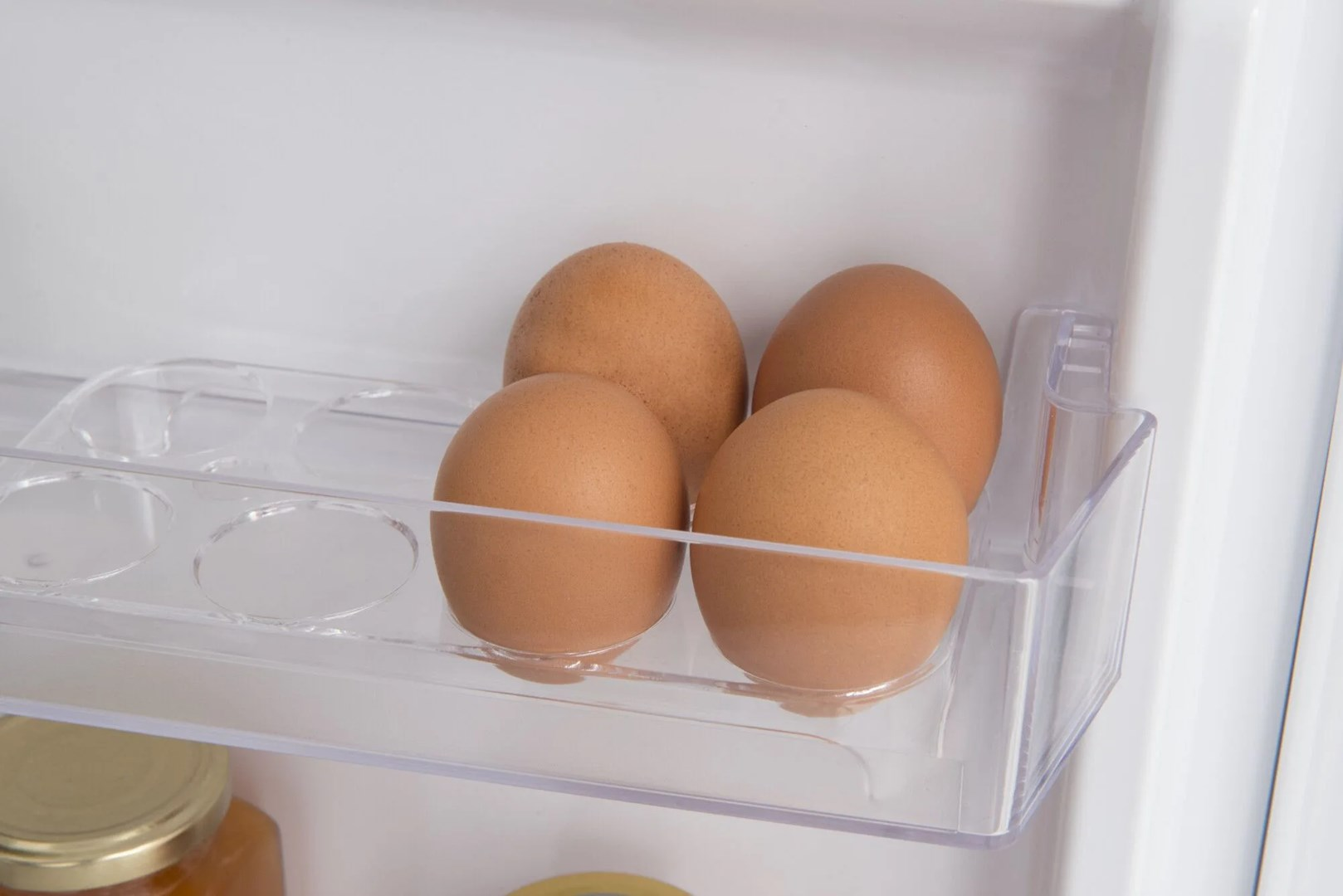 Cztery brązowe jajka w przezroczystym pojemniku na jajka w lodówce. Tło jest białe.