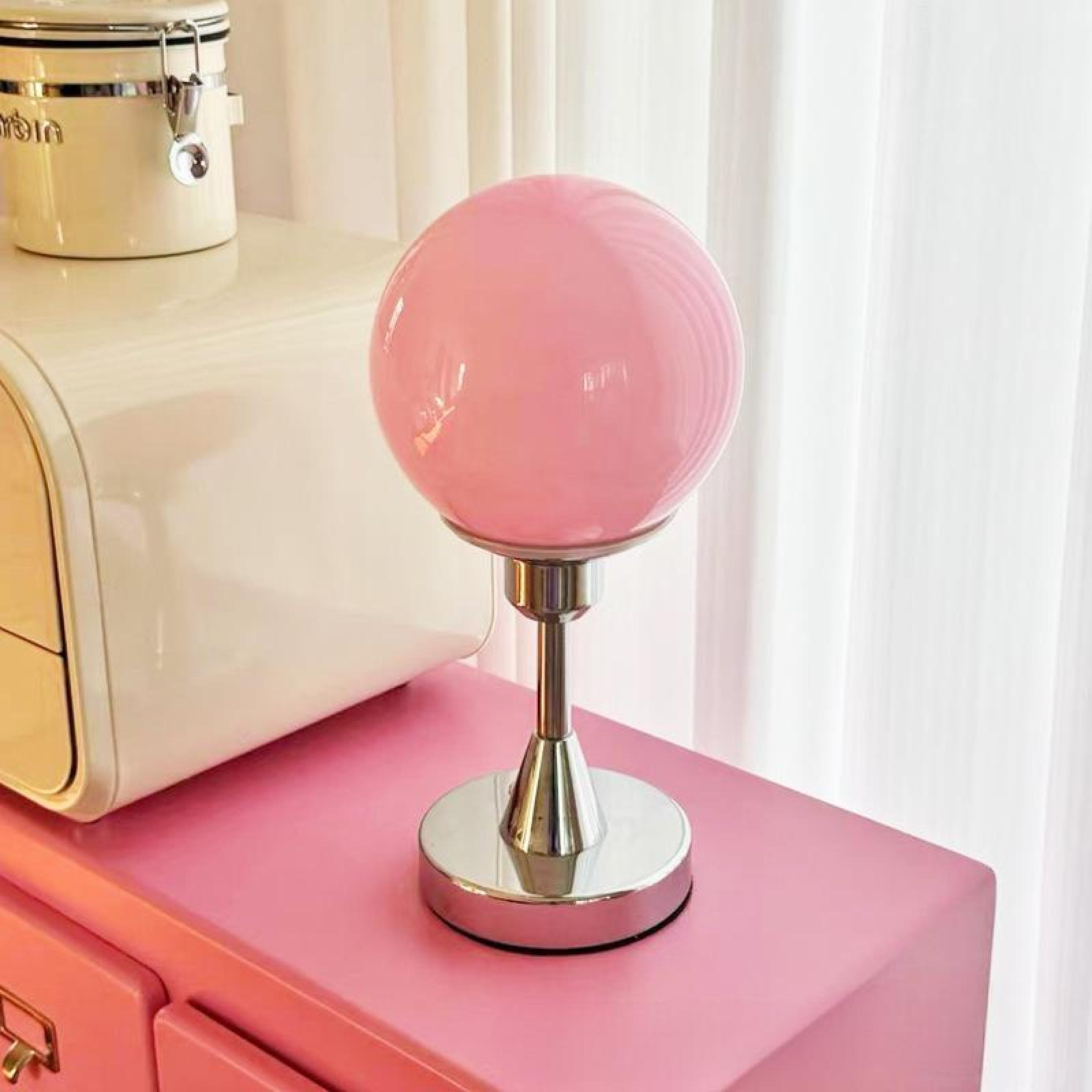 Eine rosa Kugellampe steht auf einer rosa Oberfläche. Links sind ein weißer Schrank und ein Glas. Weiße Vorhänge im Hintergrund.