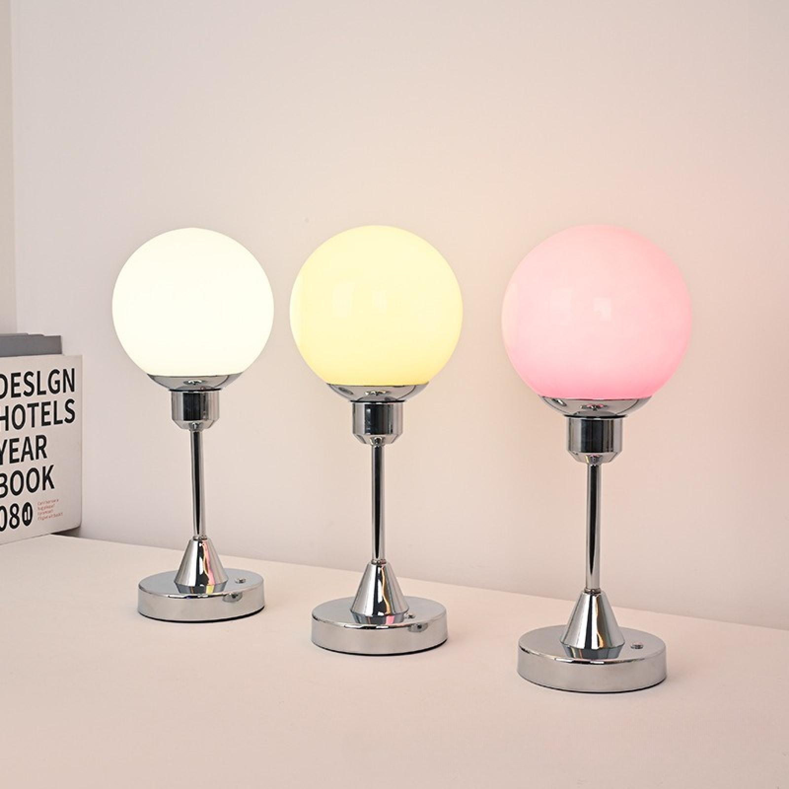 Drei Lampen mit unterschiedlich farbigen Kugeln: weiß, gelb und rosa. Sie stehen auf einer weißen Oberfläche mit einem Buch im Hintergrund.
