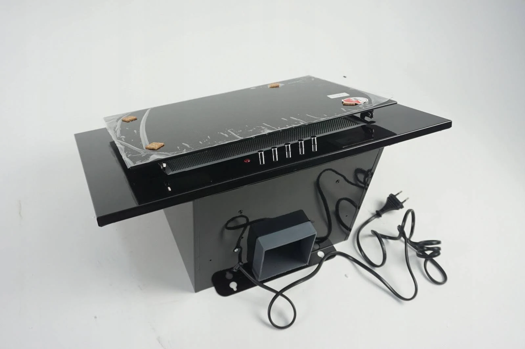 Czarny okap z folią ochronną. Posiada panel sterowania i przewód zasilający na białym tle.