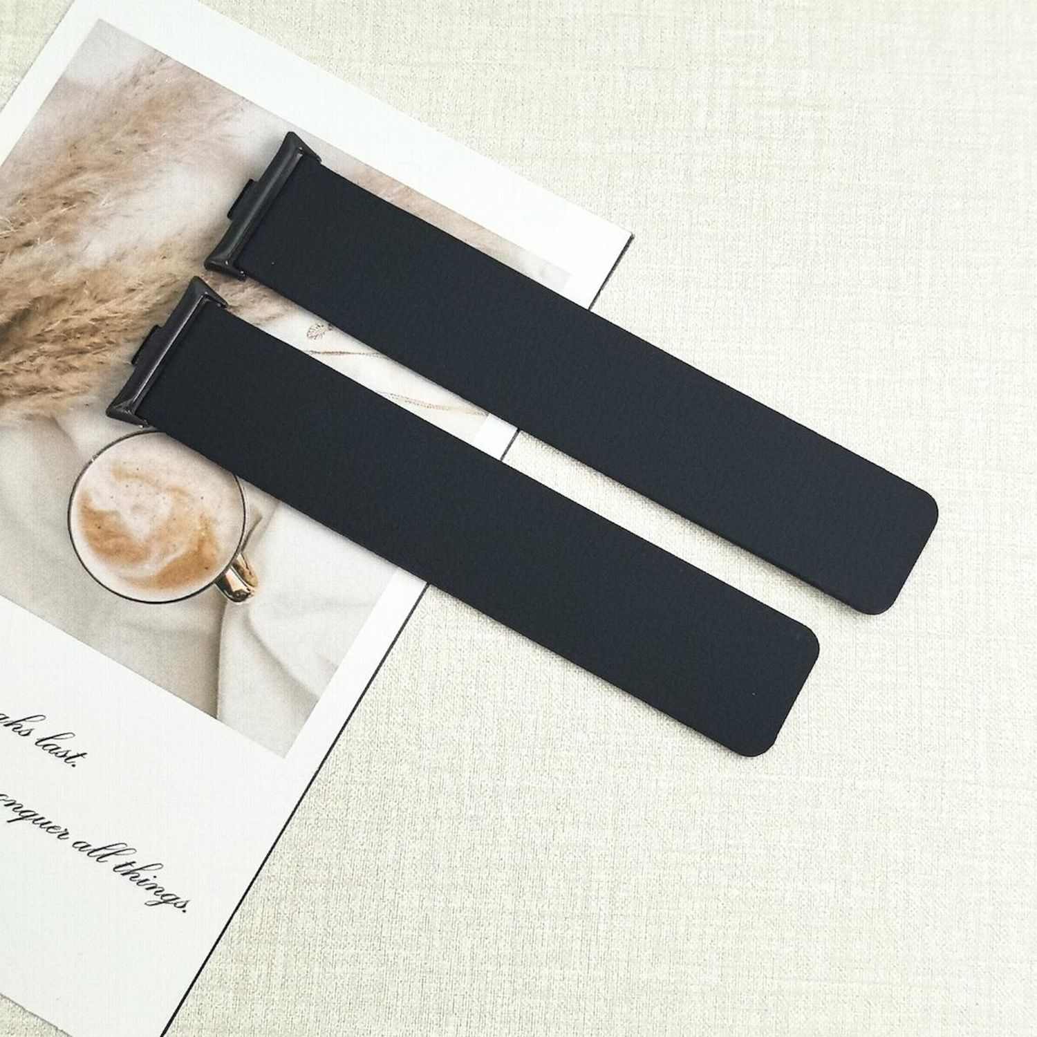 Zwei schwarze Uhrenarmbänder auf strukturierter Oberfläche, mit Kaffeetasse und Text auf Papier im Hintergrund.