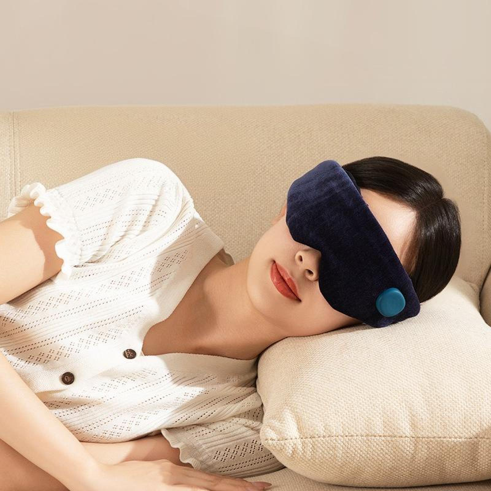 Eine Frau mit einer marineblauen Augenmaske ruht sich auf einer cremefarbenen Couch mit einem Kissen aus.
