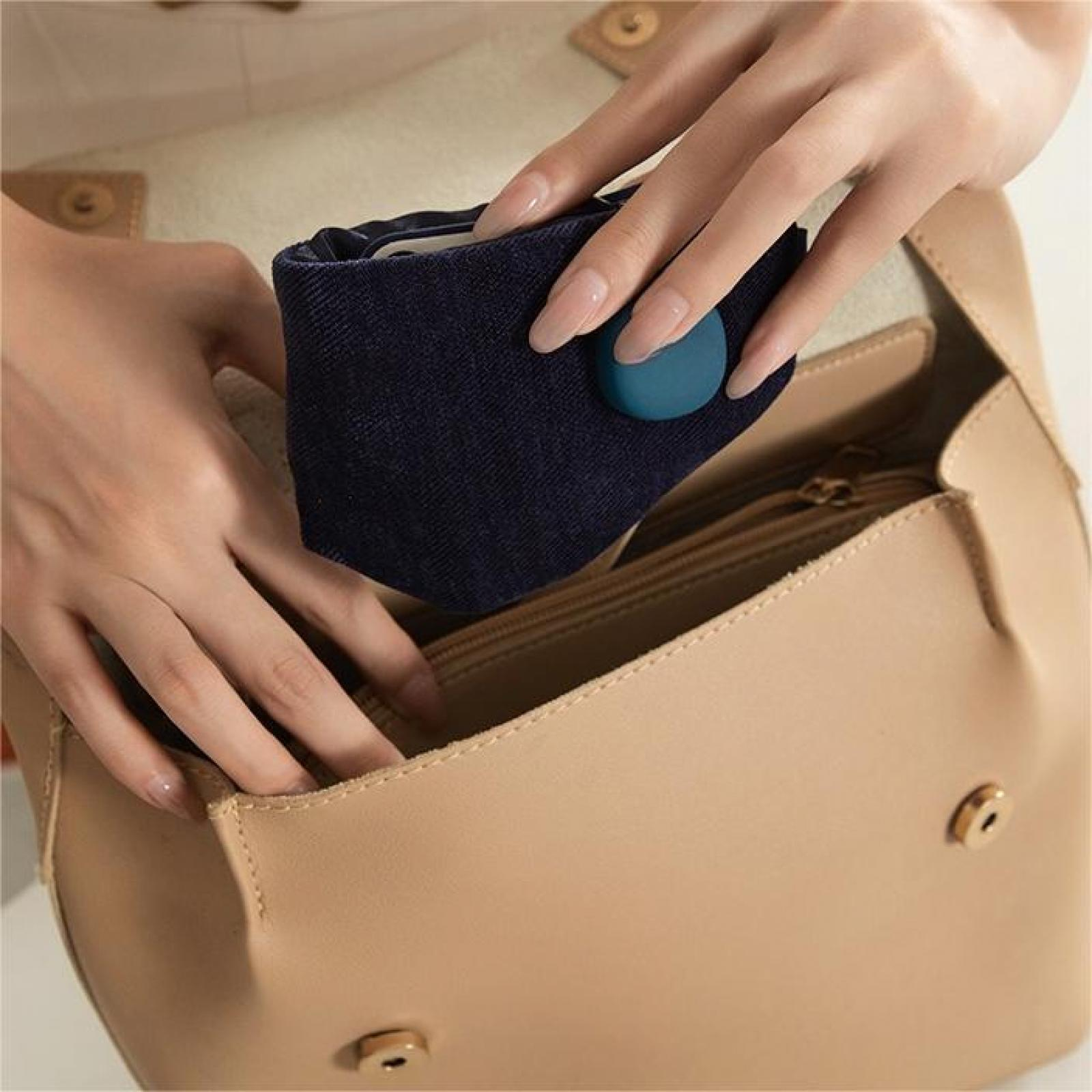 Hände legen einen marineblauen Gegenstand mit einem tealfarbenen Knopf in eine beige Handtasche mit goldfarbener Hardware.