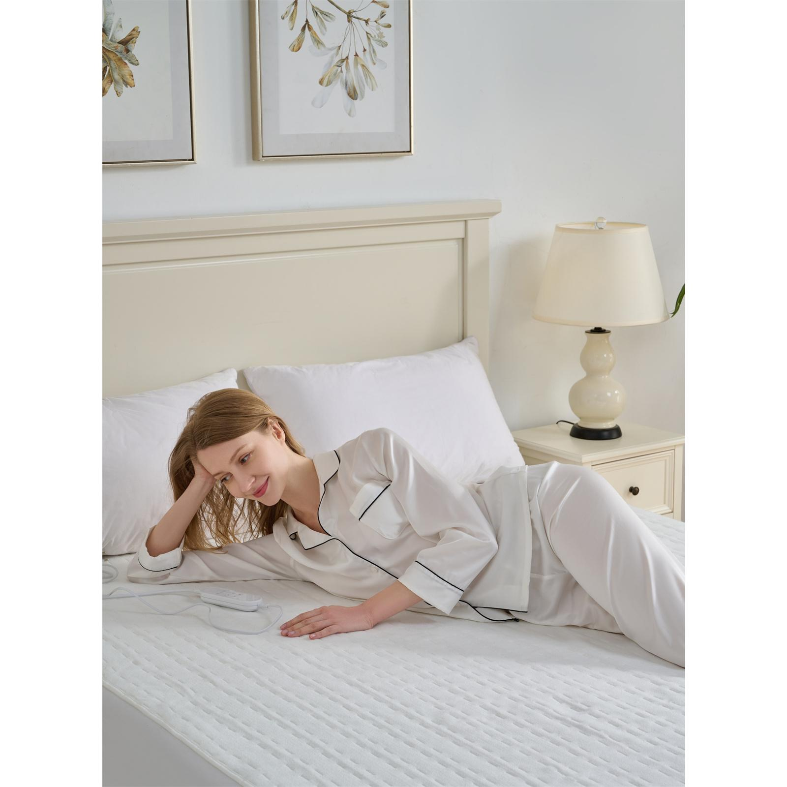 Frau in weißem Pyjama liegt auf einem Bett mit weißer Bettdecke. Es gibt eine Nachttischlampe und Wandkunst.
