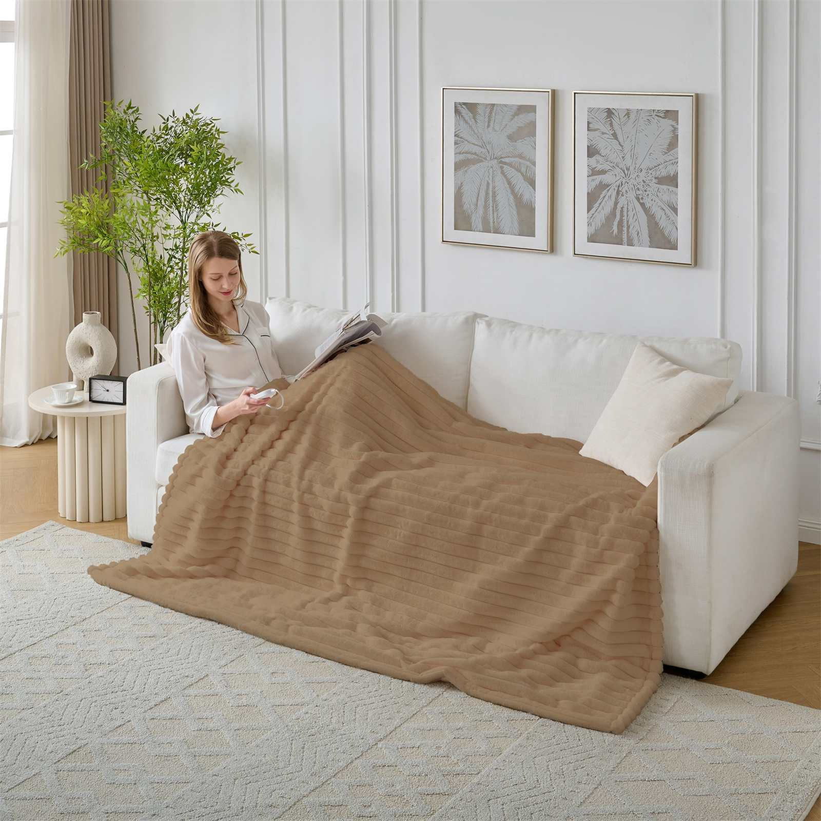 Eine Frau sitzt auf einem Sofa unter einer braunen Decke und liest. Palmenkunst hängt an der Wand. Eine grüne Pflanze befindet sich in der Nähe des Sofas.