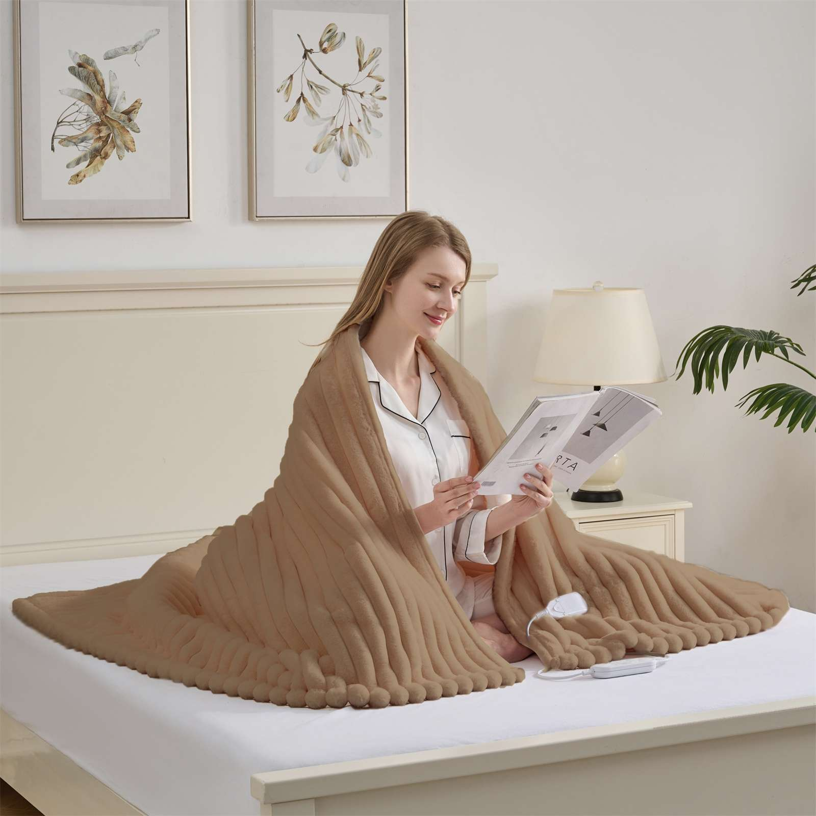 Eine Frau liest unter einer braunen Decke auf einem Bett. Über dem Bett hängen zwei gerahmte Bilder. Eine Lampe und eine Pflanze sind im Hintergrund.