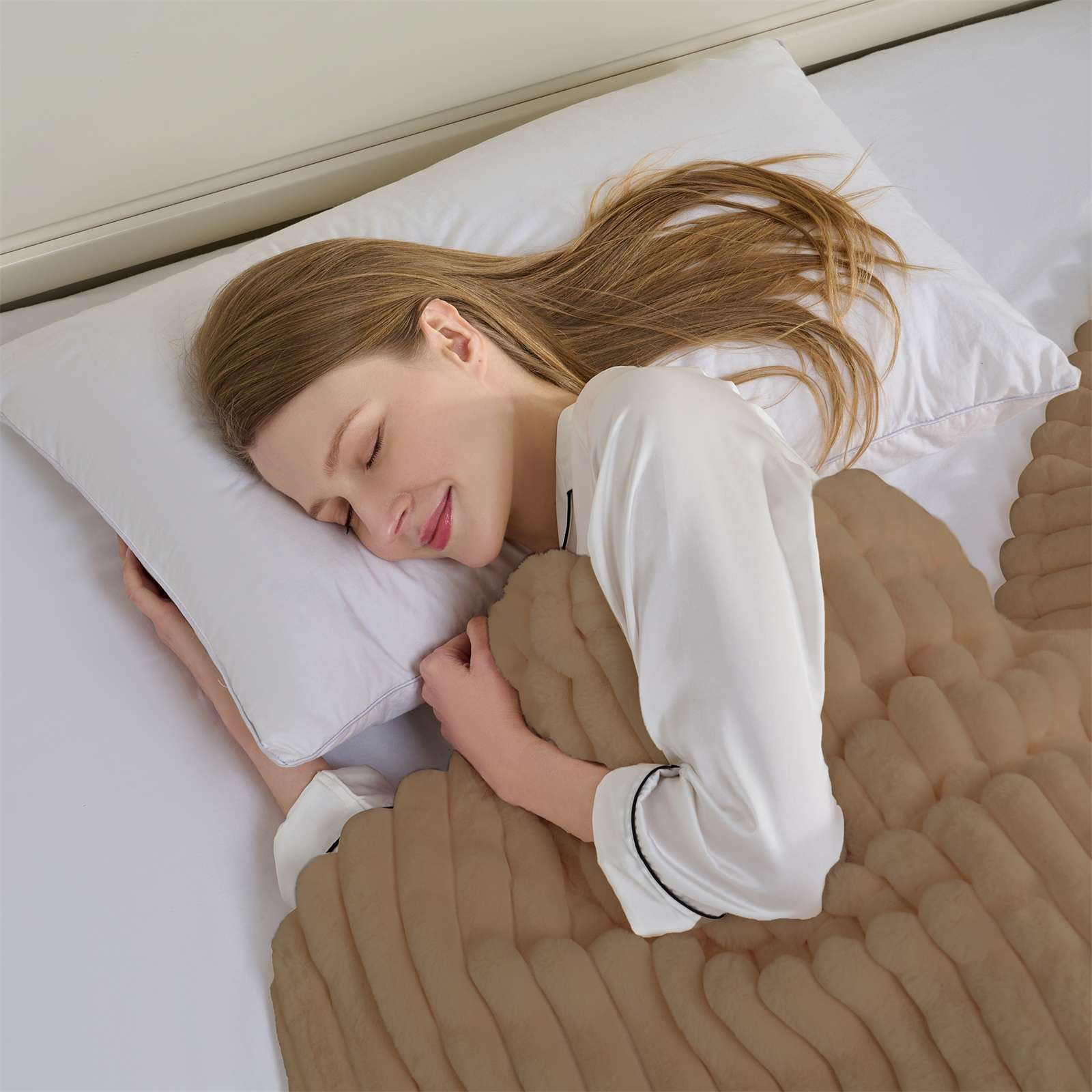 Eine Frau schläft im Bett, bedeckt von einer braunen Decke. Sie trägt ein weißes Oberteil. Unter ihrem Kopf liegt ein weißes Kissen.