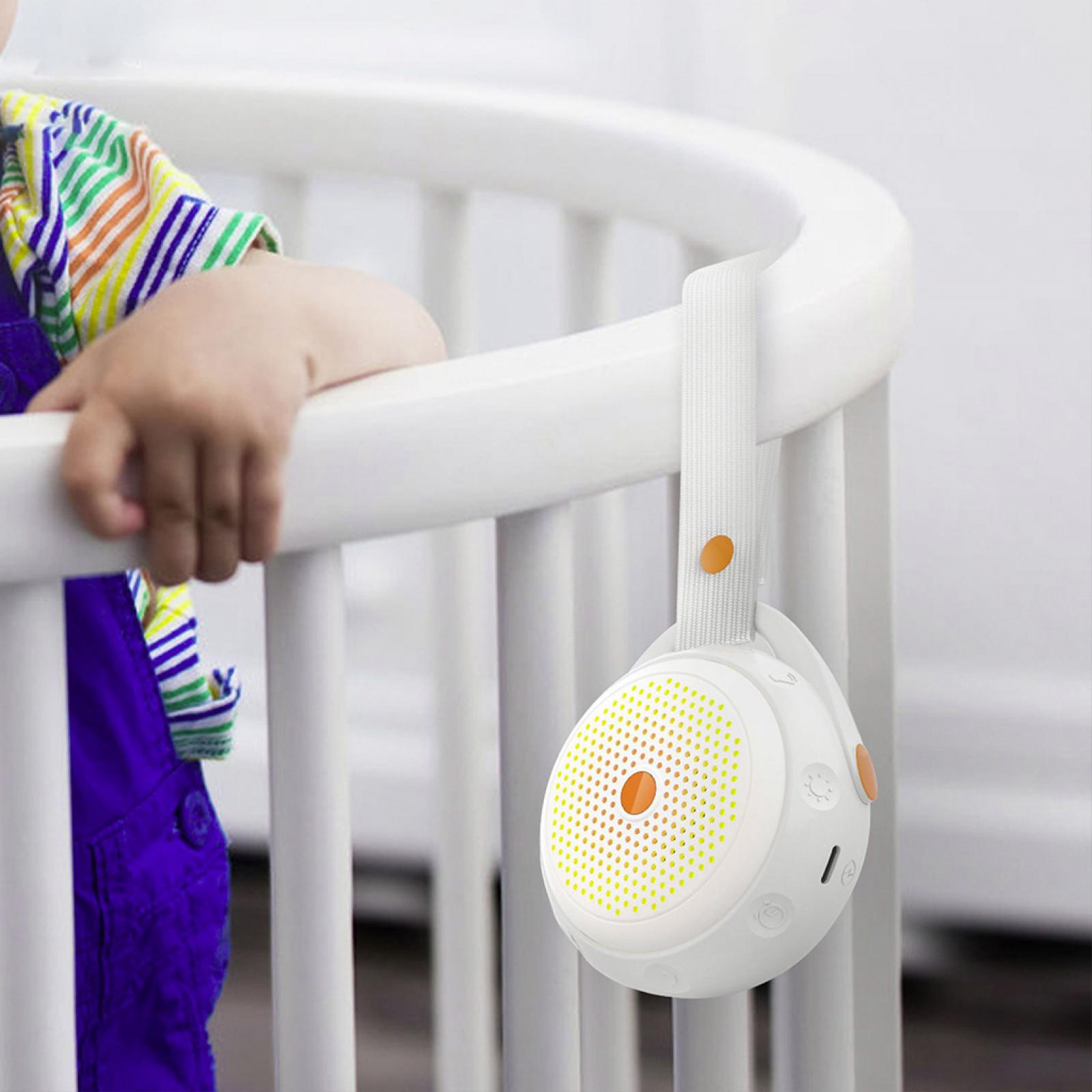 Die Hand eines Babys liegt auf einem weißen Kinderbett. Ein weißes Soundgerät mit orangefarbenen Akzenten hängt am Bett.