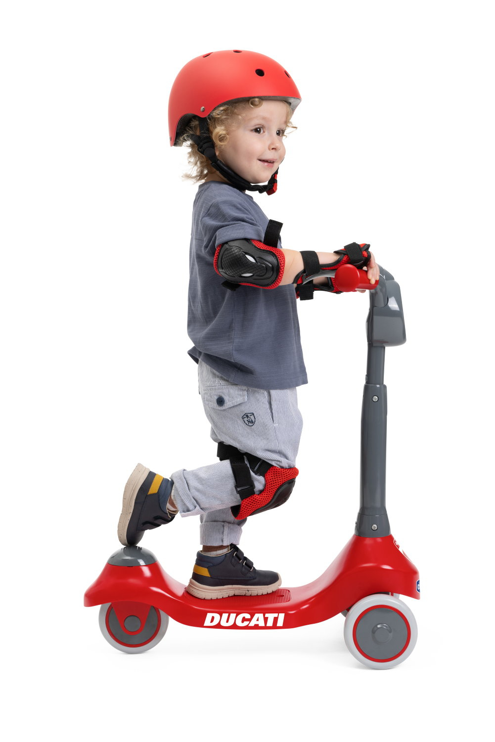 Ein Kind mit rotem Helm und Schutzkleidung fährt einen roten Roller. Auf dem Roller steht 'DUCATI'.