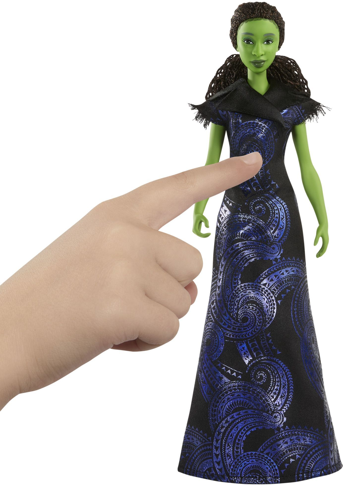 Una muñeca de piel verde con un vestido negro y azul, con un dedo apuntando hacia ella. La muñeca tiene cabello castaño y está sobre fondo blanco.