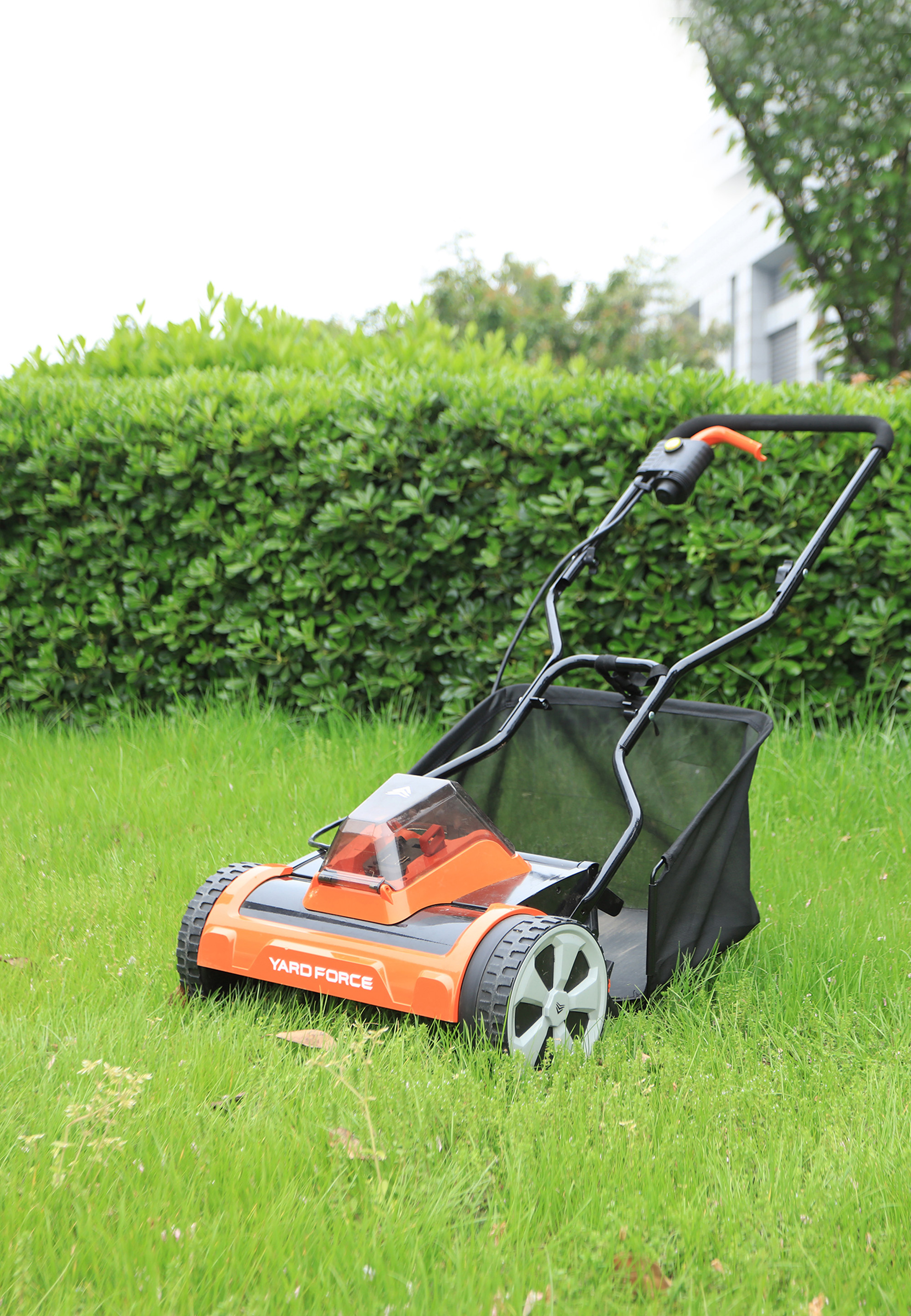 Ein orangefarbener und schwarzer Rasenmäher auf grünem Gras. Im Hintergrund sind eine Hecke und ein Gebäude zu sehen.