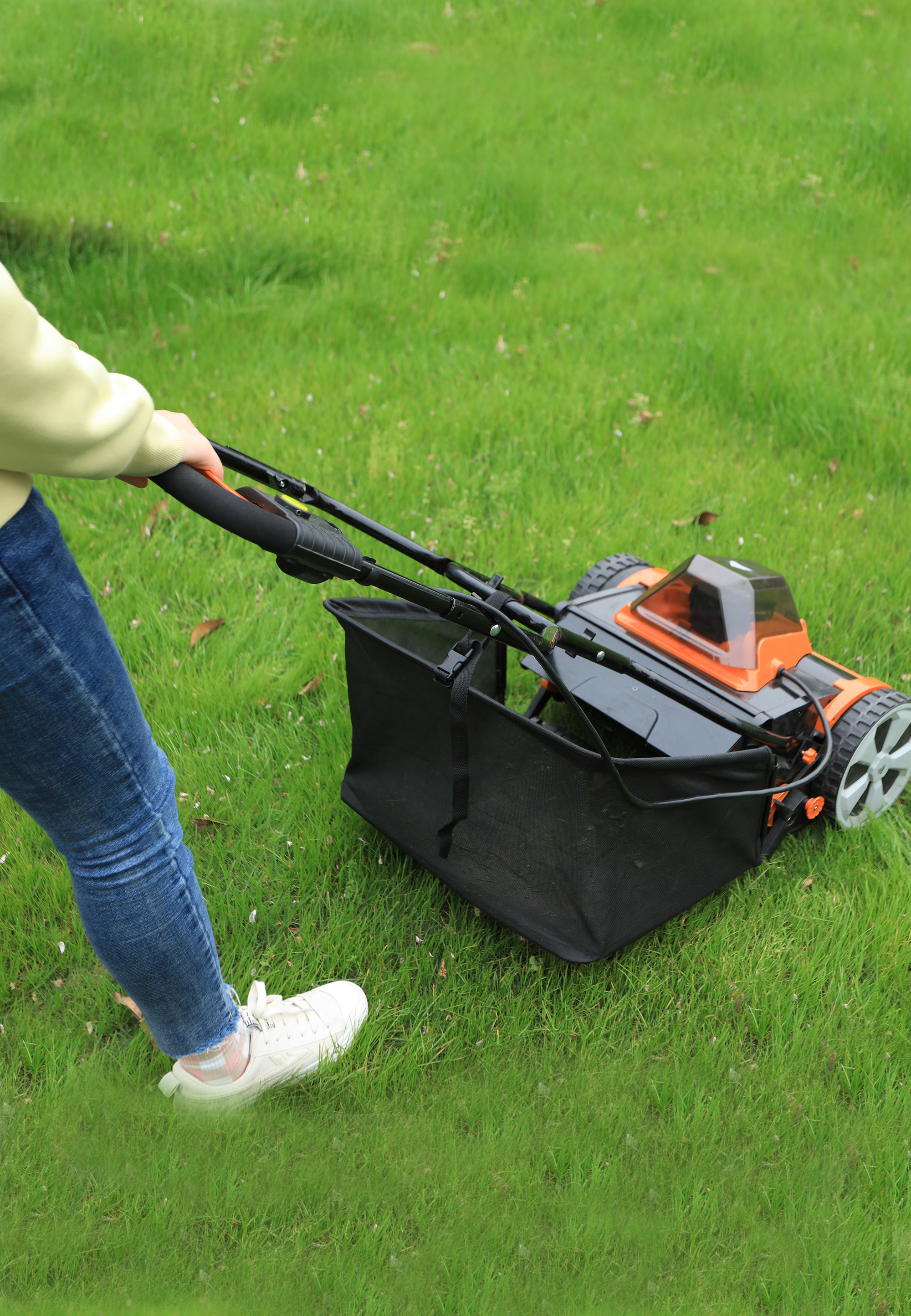 Eine Person mäht den grünen Rasen mit einem schwarz-orangenen Rasenmäher. Die Person trägt blaue Jeans und weiße Schuhe.