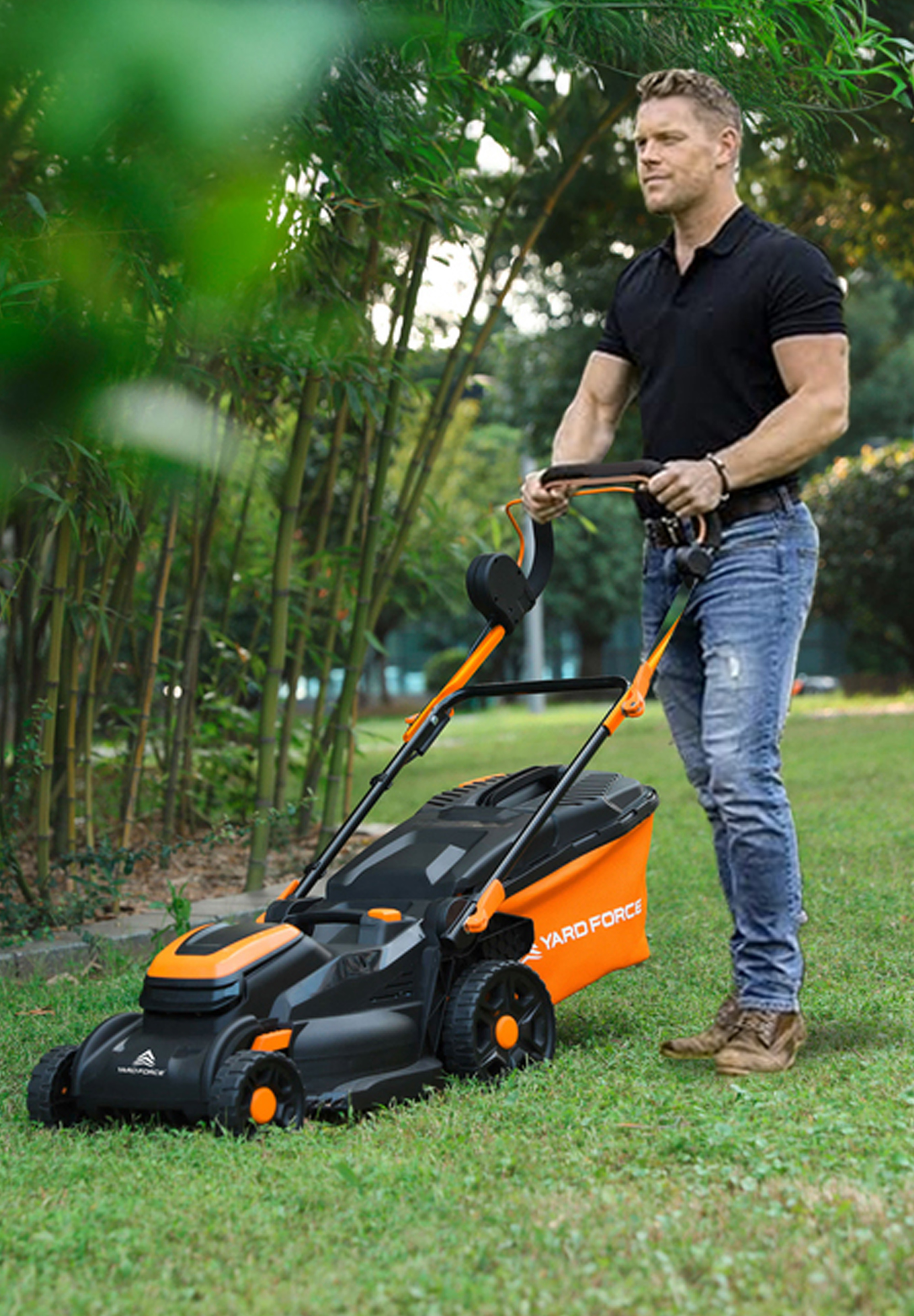 Ein Mann in einem schwarzen Hemd und Jeans schiebt einen Rasenmäher auf Gras. Der Mäher ist orange und schwarz, mit einem Beutel.
