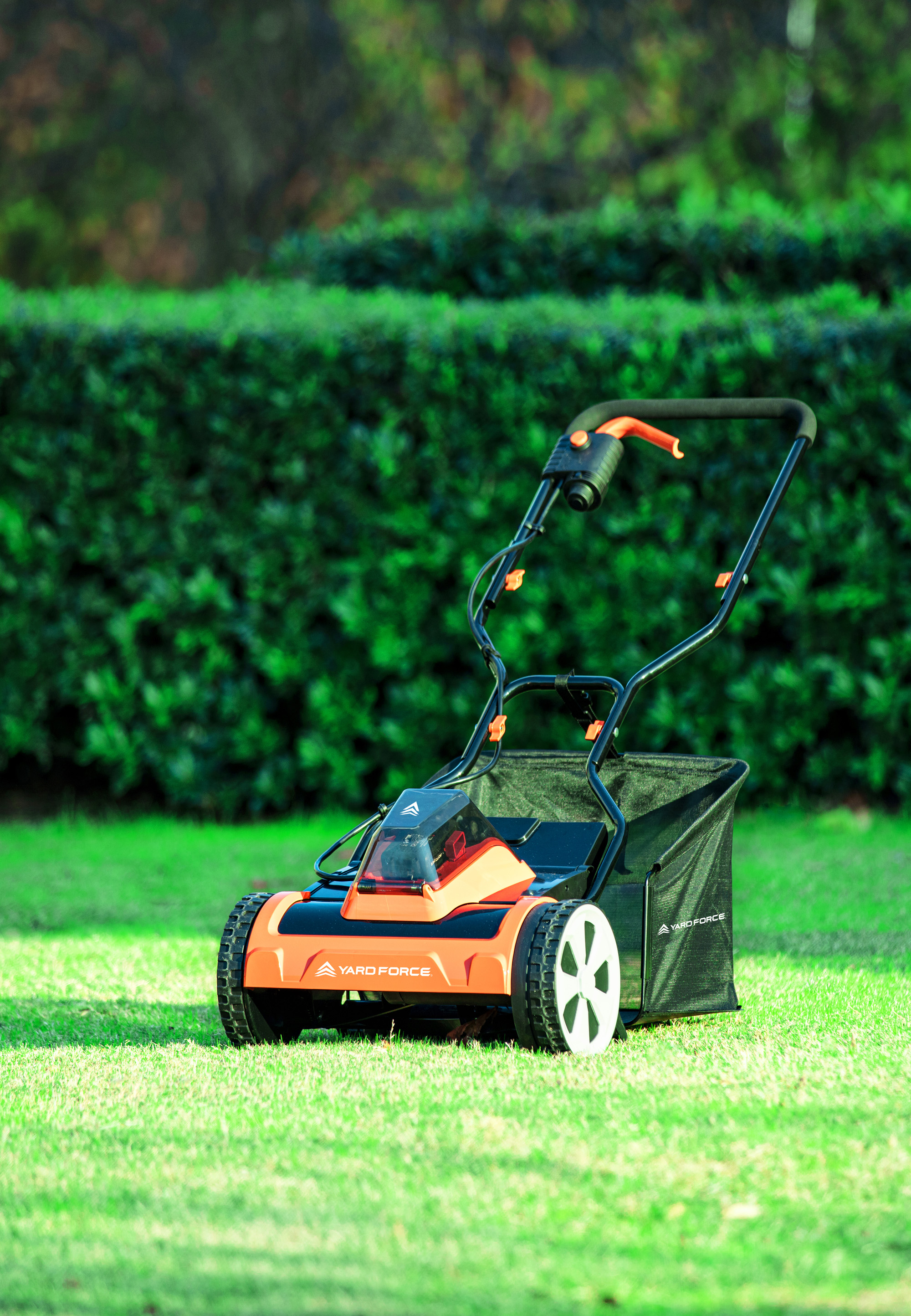 Ein orangefarbener und schwarzer Rasenmäher auf grünem Gras. Im Hintergrund ist eine grüne Hecke zu sehen.