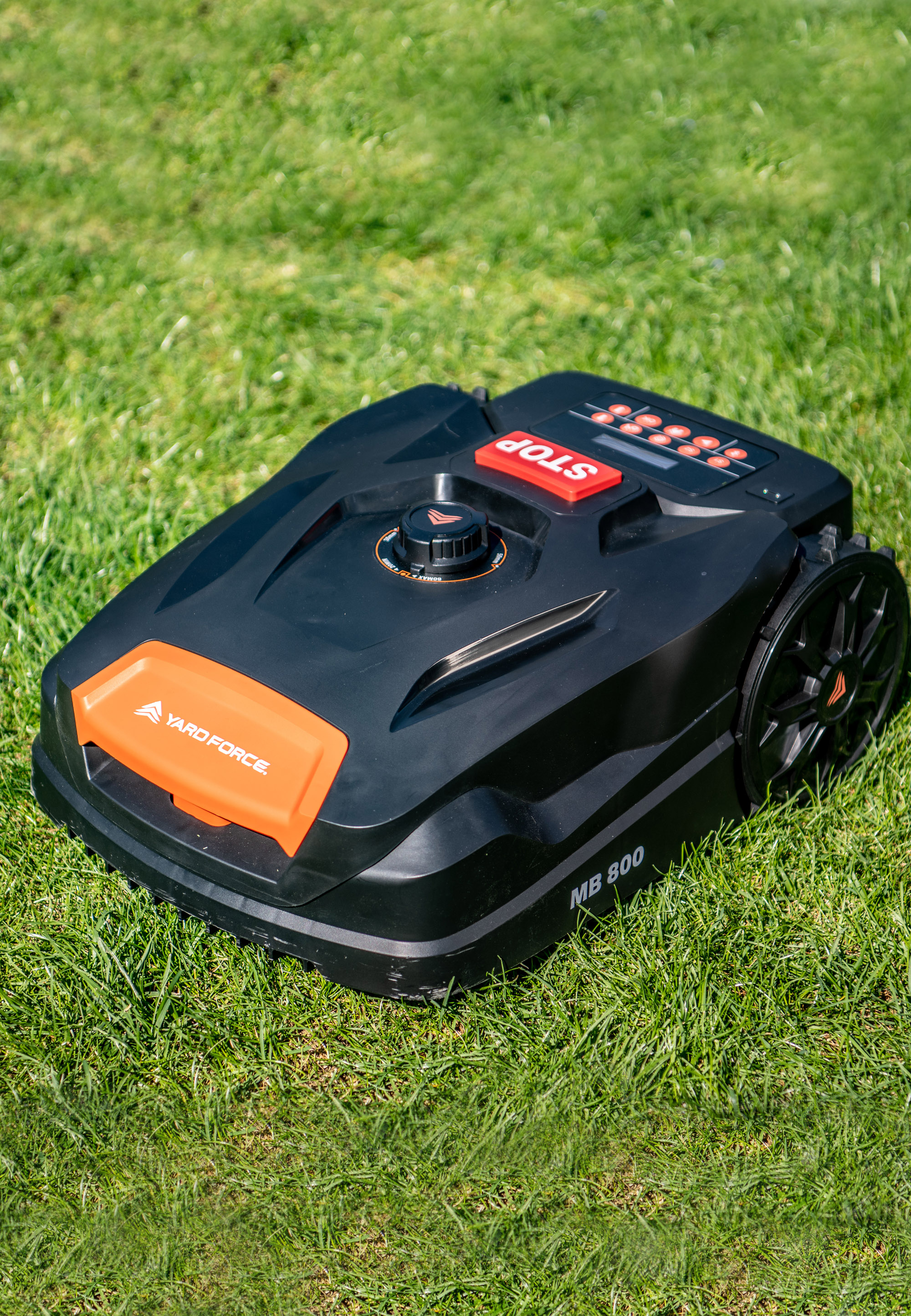 Schwarzer Yard Force MB 800 Mähroboter auf grünem Gras. Enthält orangefarbene Akzente und eine rote STOP-Taste.