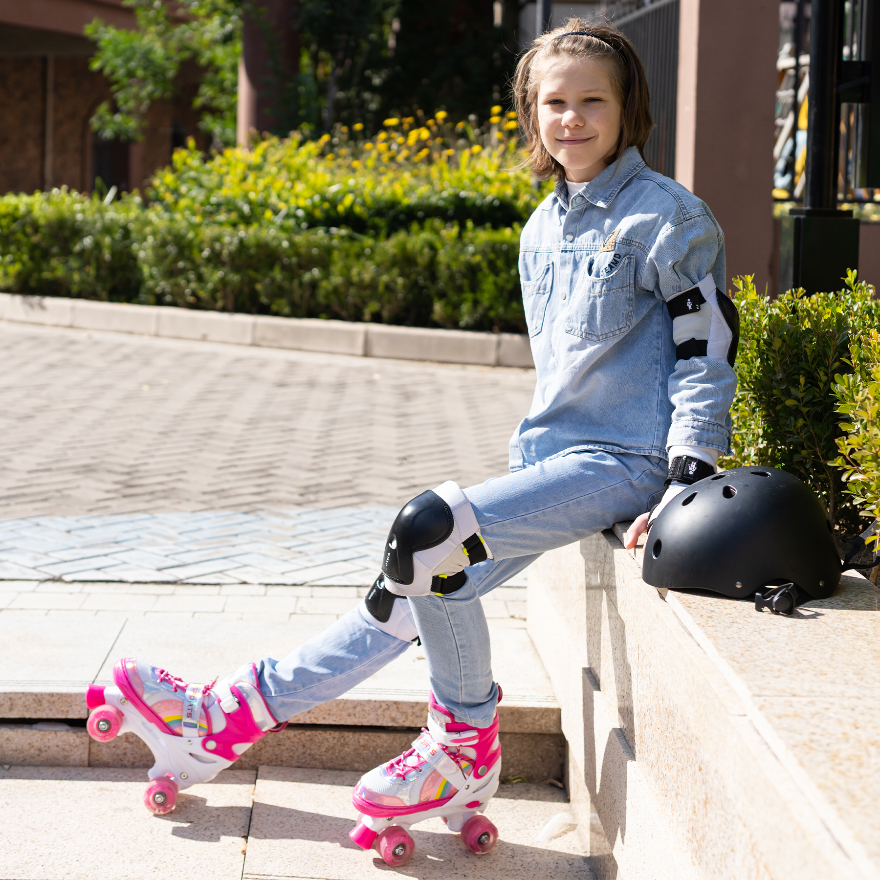 Ein Mädchen in Jeans und Jeanshemd sitzt mit Rollschuhen. Ein schwarzer Helm und Schutzkleidung sind vorhanden.