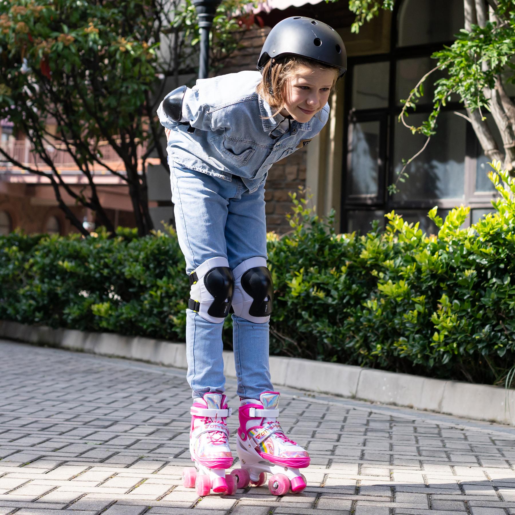 Ein Mädchen in Jeans, Helm und Schutzkleidung steht auf rosa Rollschuhen. Grüne Büsche und ein Backsteingebäude sind im Hintergrund.
