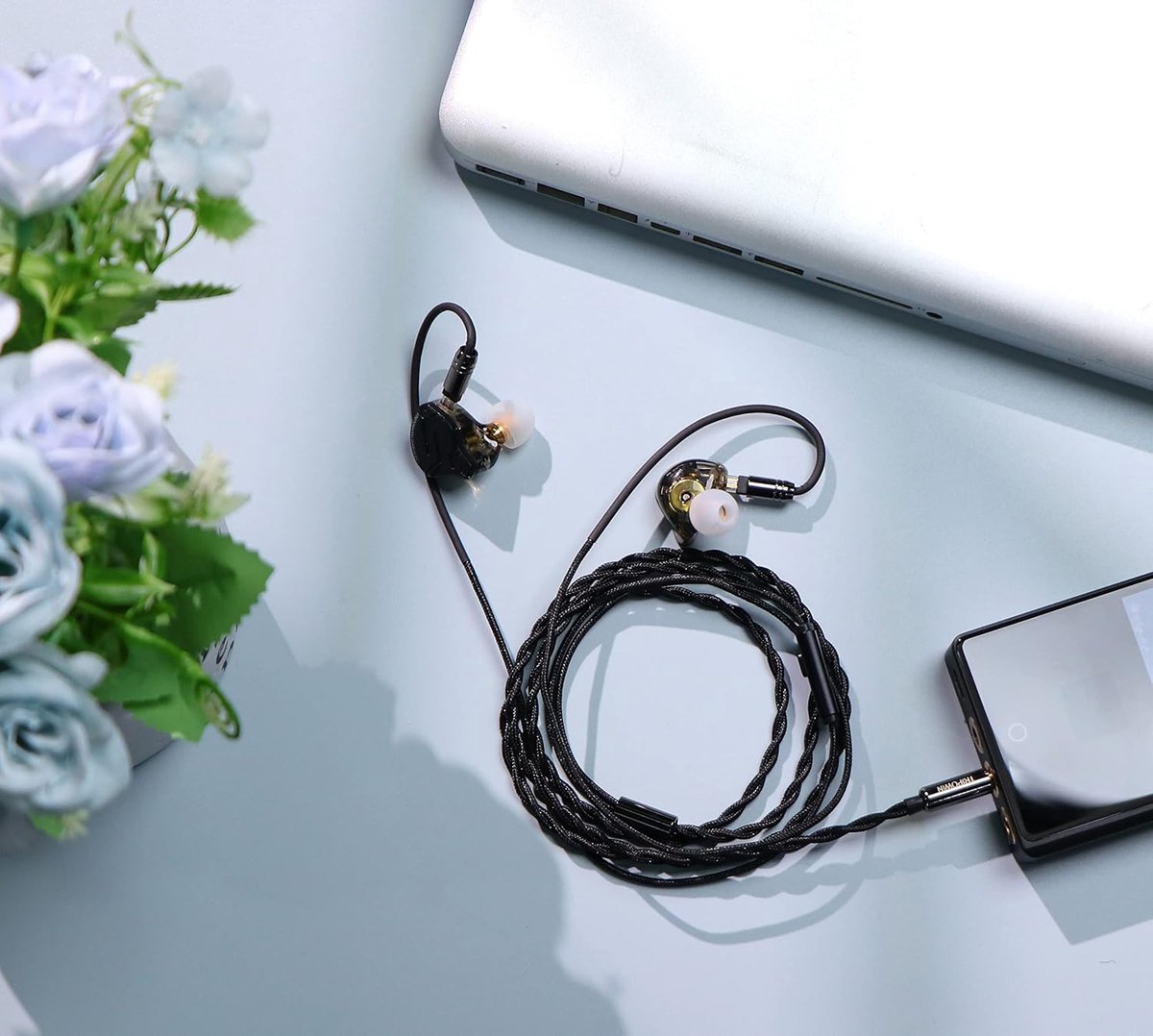 Auricolari con cavo intrecciato nero, collegati a uno smartphone. Laptop e fiori sono anche nella scena.