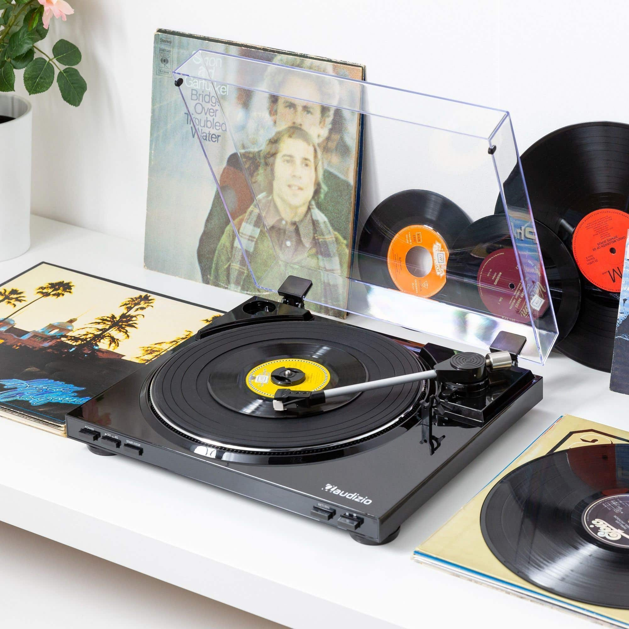 A turntable with a record playing. Records and album covers are in the background. A pink flower and green leaves are in a pot.