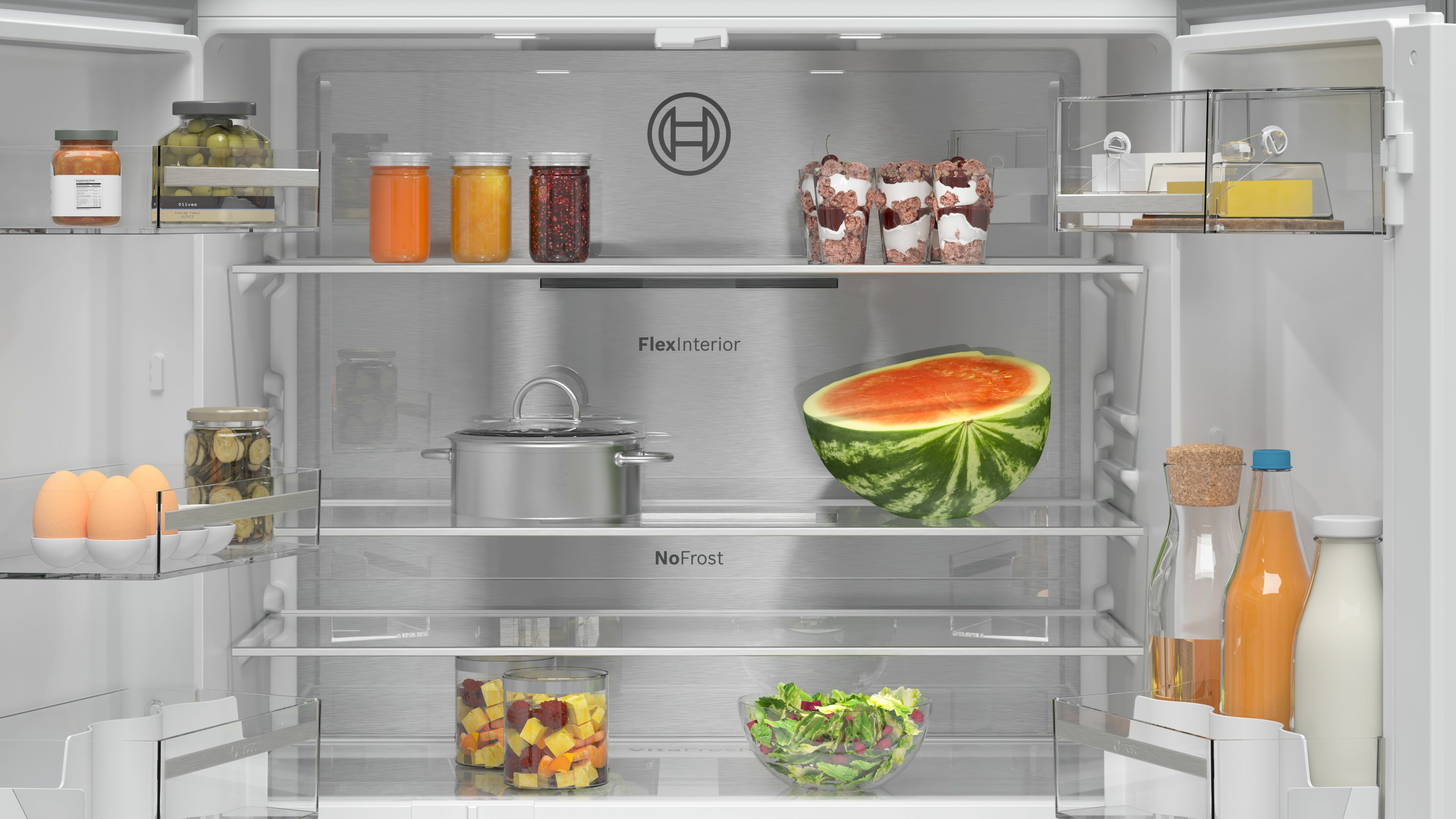 Interieur van een koelkast met diverse etenswaren: watermeloen, potten jam, salade, eieren, sap en andere items.