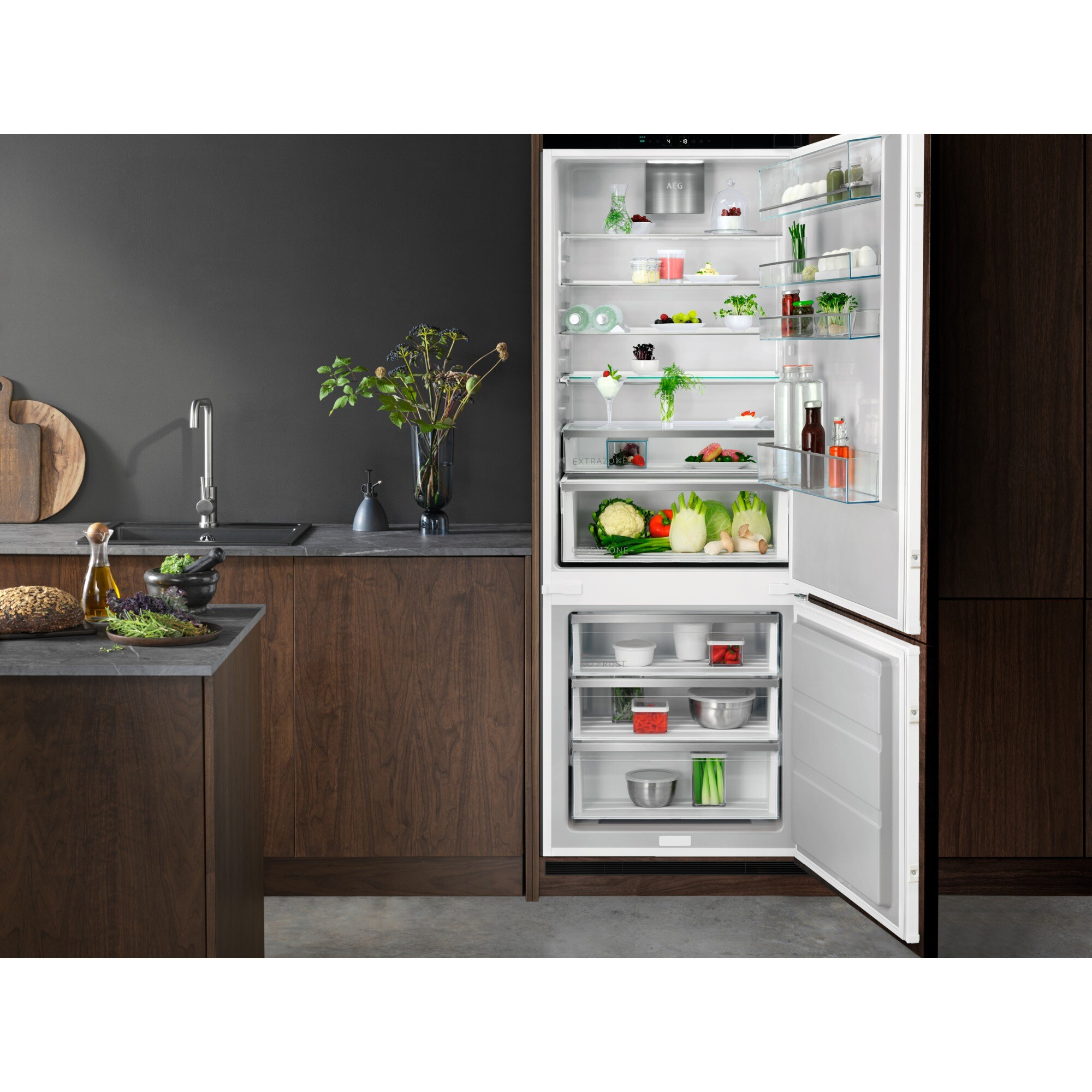 Una cocina con nevera abierta llena de comida. Encimera con ingredientes. Gabinetes de madera oscura y pared gris.