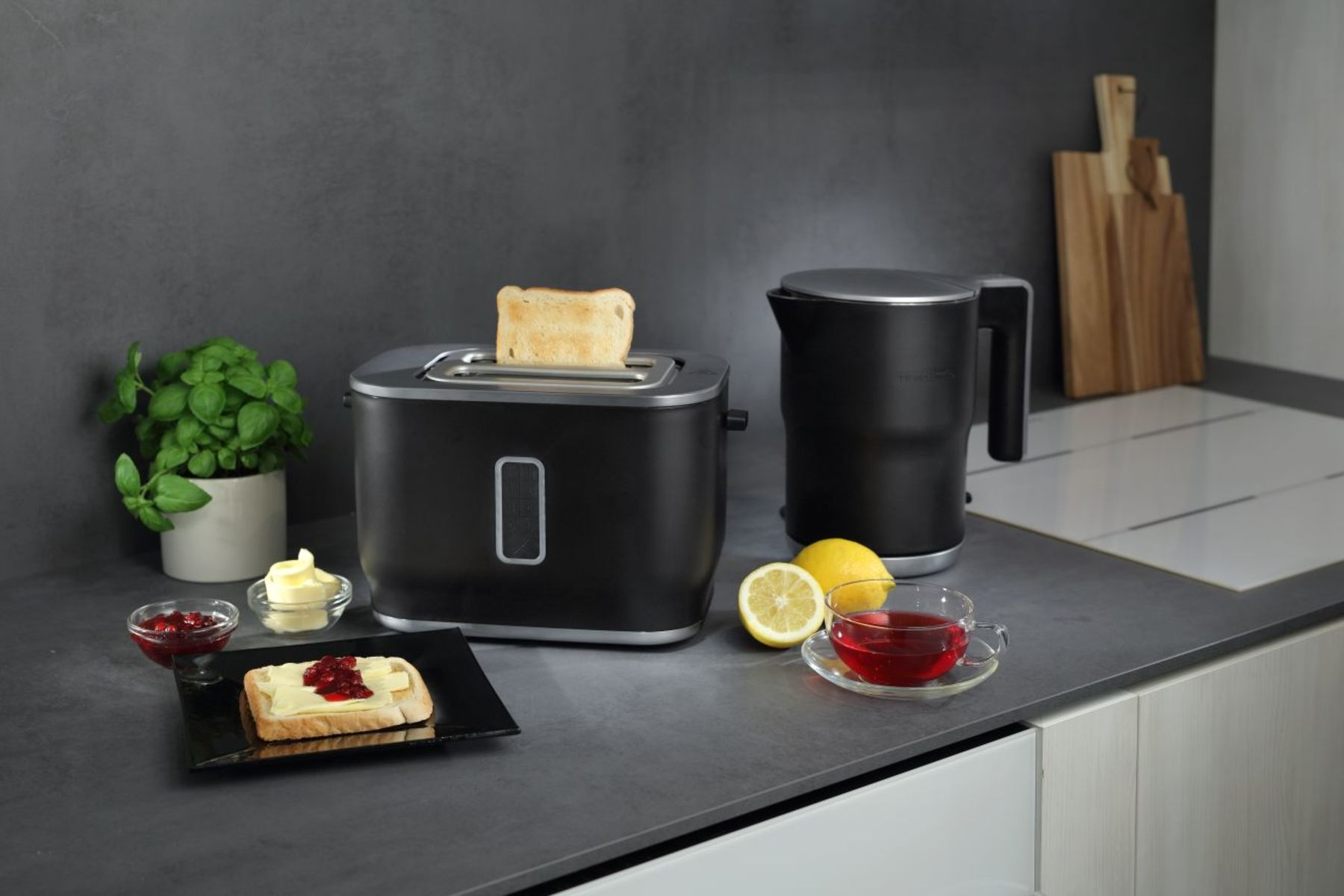 Eine Küchenszene mit Toaster, Wasserkocher, Toast mit Marmelade, Tee, Zitrone und einer Pflanze. Graue Theke und Hintergrund.