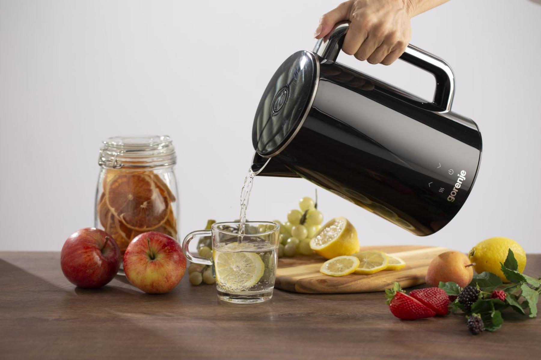Eine Hand gießt Wasser aus einem schwarzen Wasserkocher in ein Glas Zitronenwasser. Äpfel, Obst und ein Glas Orangen stehen auf dem Tisch.