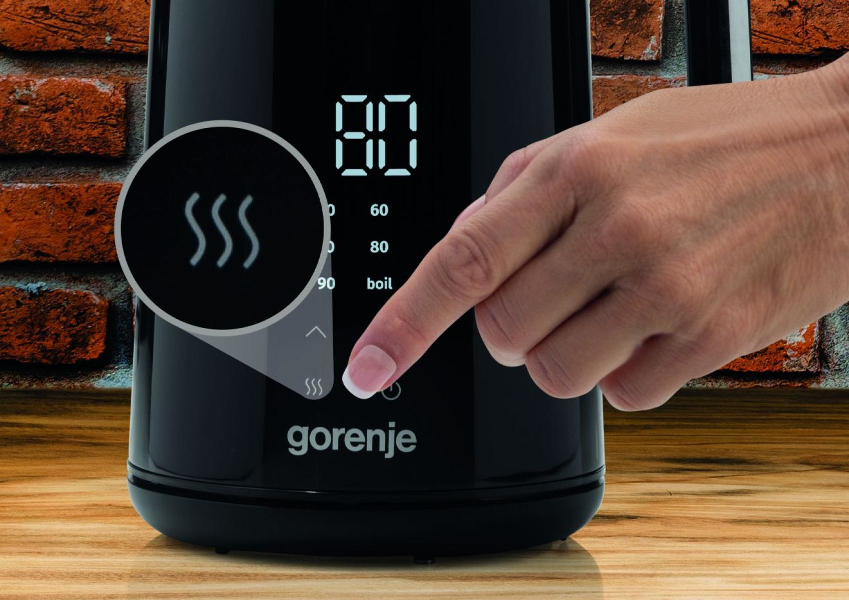 Eine Hand drückt eine Taste auf einem schwarzen Wasserkocher. Das Display zeigt '80'. Ein Dampfsymbol wird hervorgehoben. Ziegelmauer im Hintergrund.