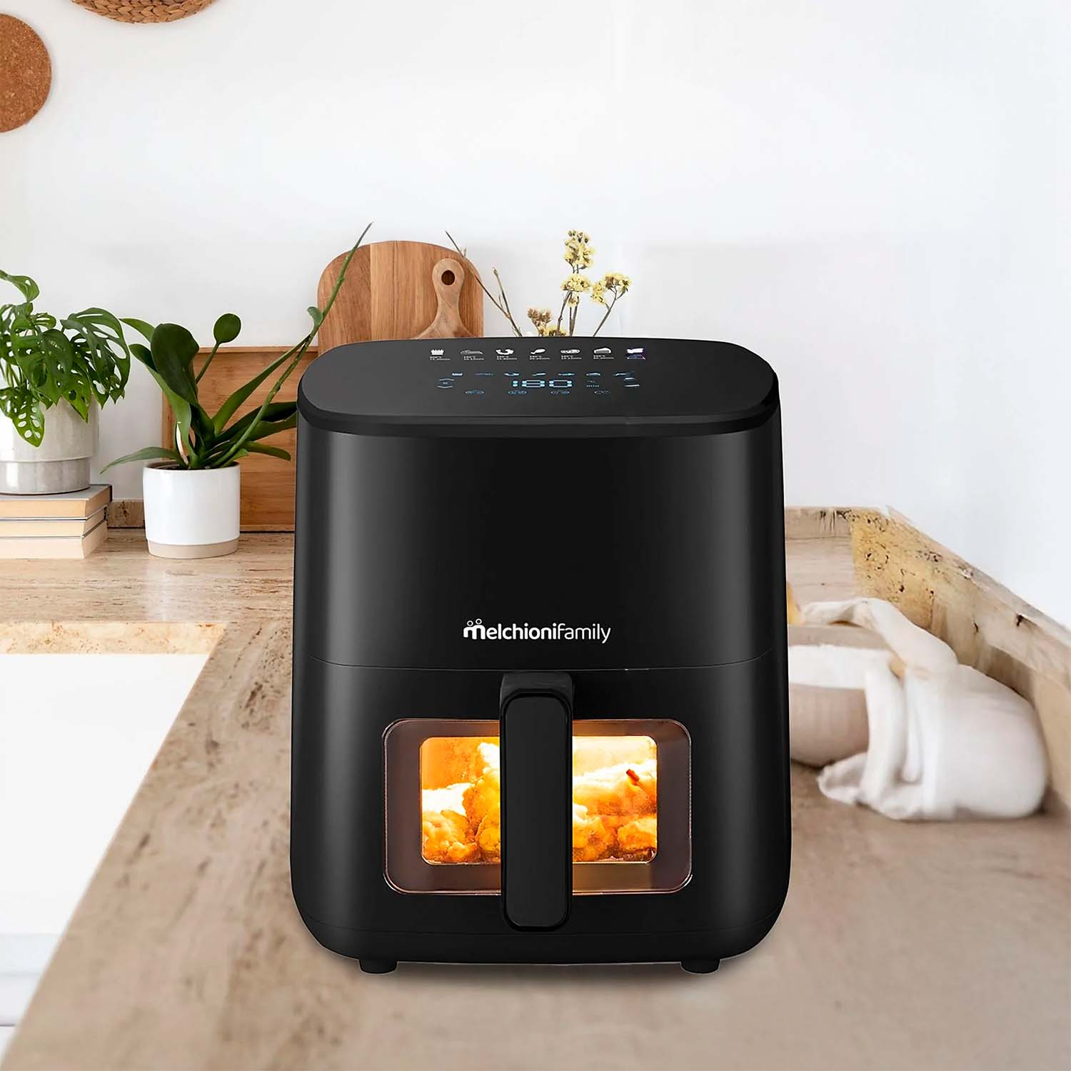 Ein schwarzer Airfryer mit Griff steht auf einer Arbeitsplatte. Im Inneren wird Essen gekocht. Pflanzen, Schneidebretter und eine weiße Wand sind im Hintergrund.
