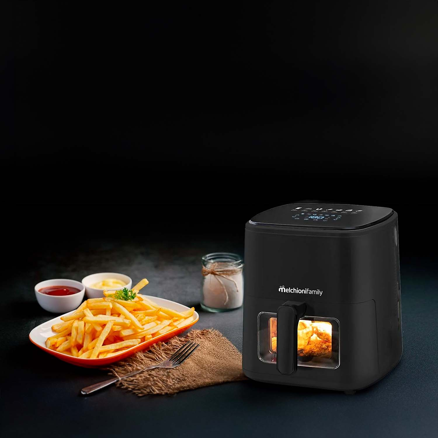 Una freidora de aire con papas fritas, salsas y sal sobre una superficie oscura. La freidora es negra con asa y vista interior.