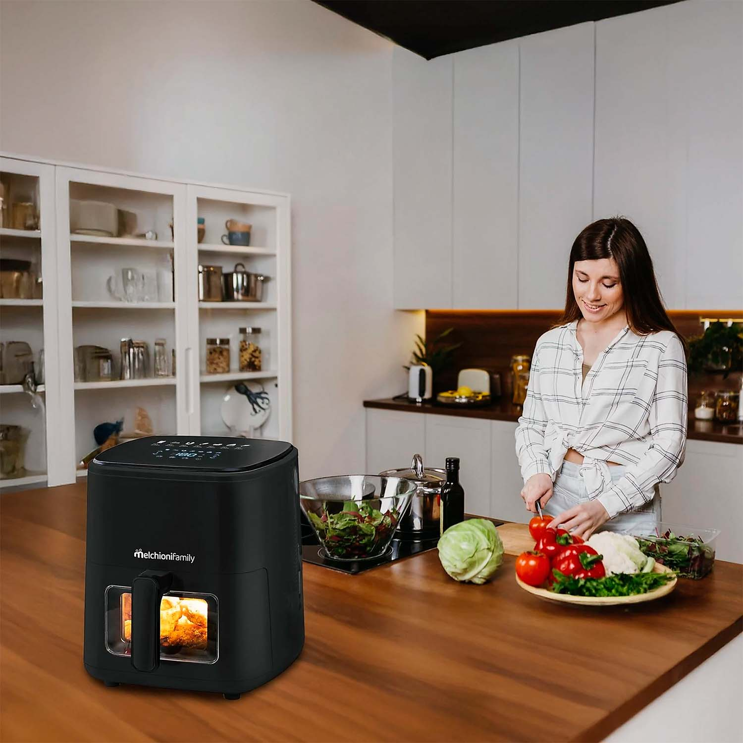 Una mujer corta tomates en una cocina. Una freidora de aire está en la encimera. También hay verduras y un bol de ensalada.