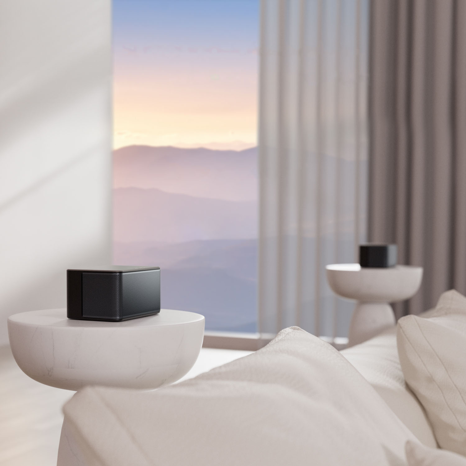 Czarny głośnik stoi na białym stoliku w pokoju. Przez duże okno widać góry. Widoczne są poduszki i drugi głośnik.