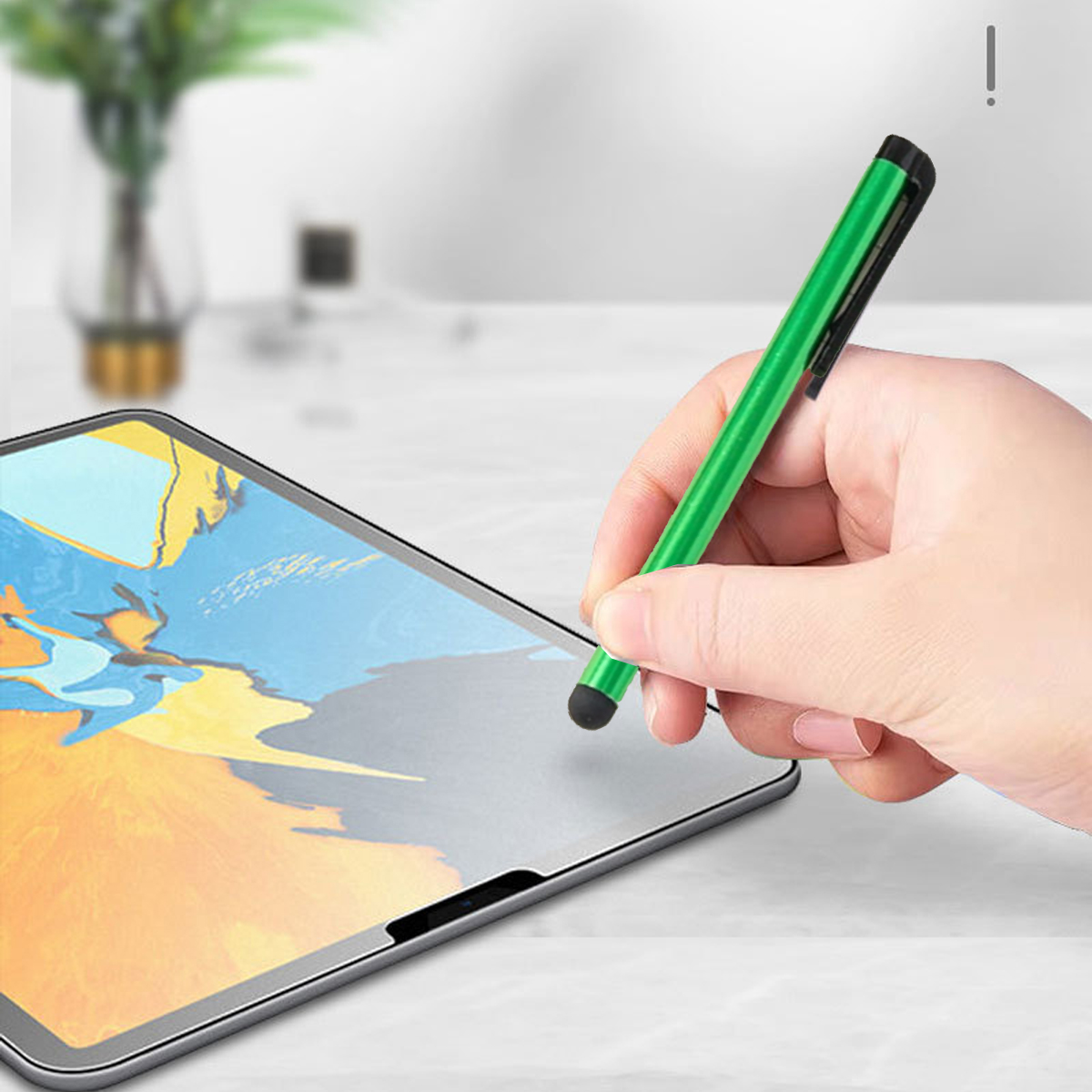 Eine Hand hält einen grünen Stift über einem Tablet. Das Tablet zeigt ein farbenfrohes Bild. Im Hintergrund ist eine Vase mit Pflanzen zu sehen.