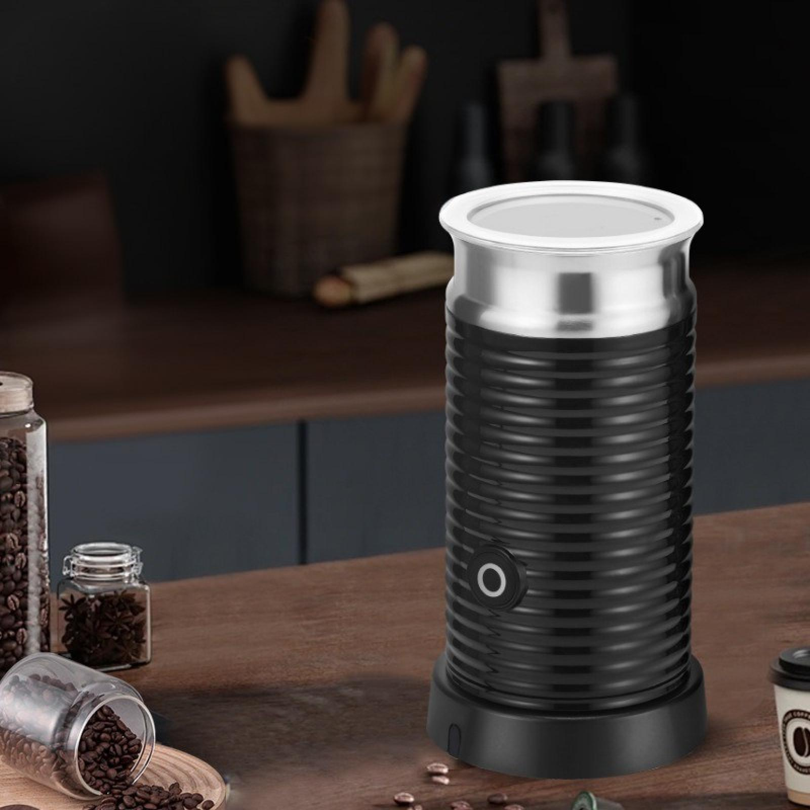 Ein schwarzer und silberner Milchaufschäumer auf einer Holzoberfläche mit Kaffeebohnen und Gläsern. Der Hintergrund ist eine Küche.