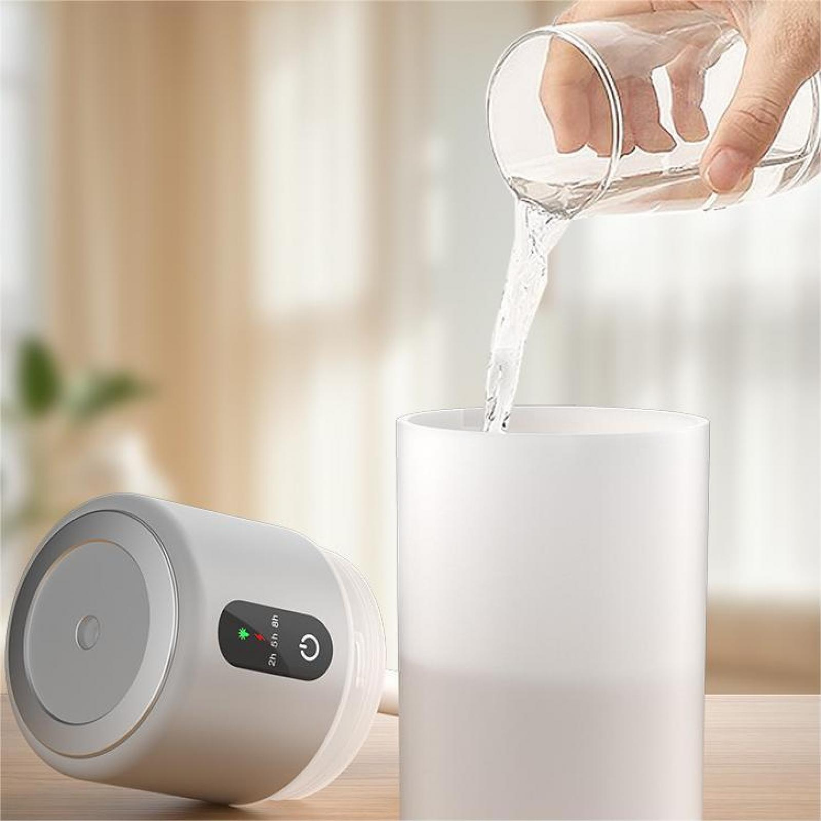 Eine Hand gießt Wasser aus einem Glas in einen weißen Zylinder. Links befindet sich ein grauer Luftbefeuchter.