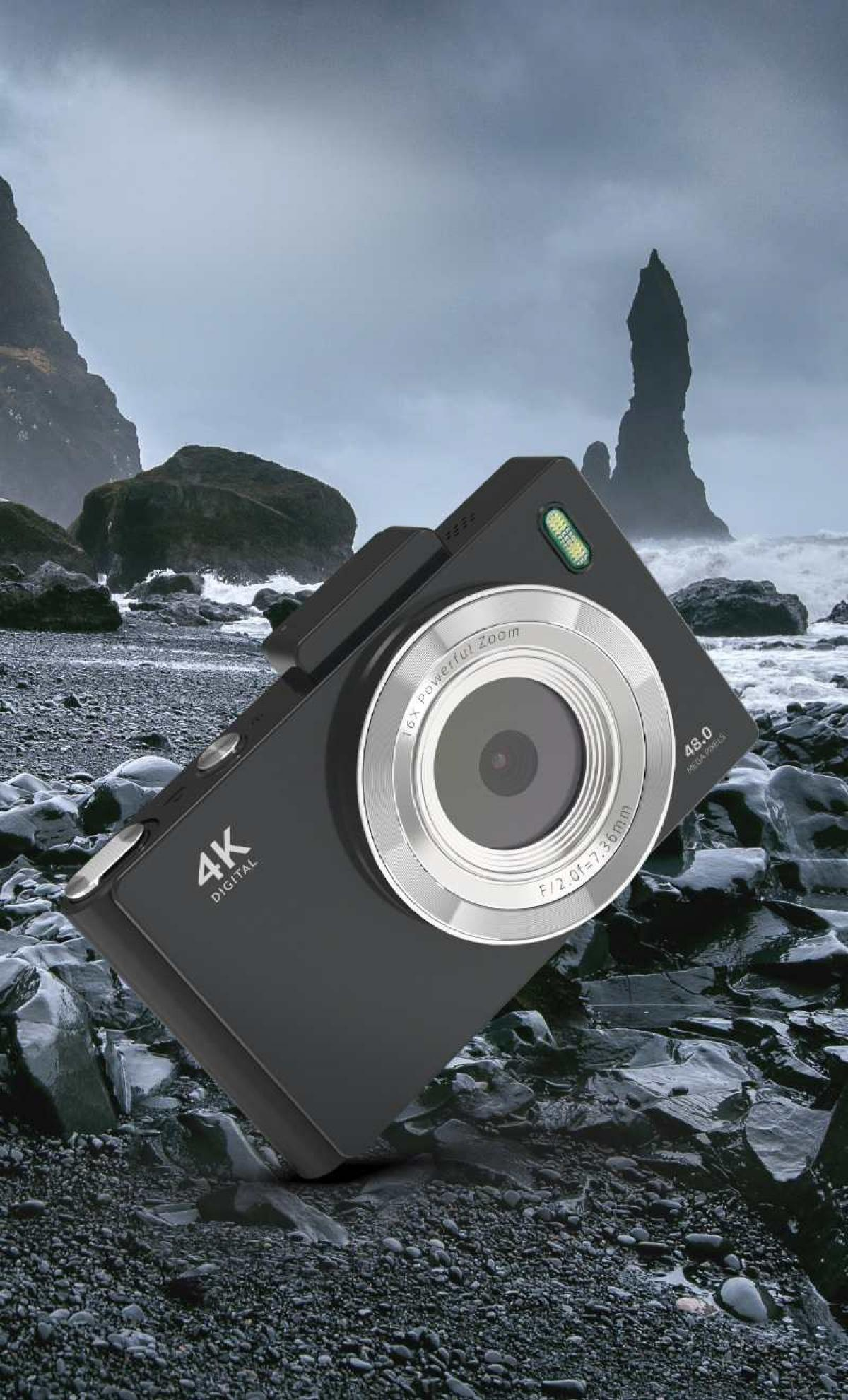 Schwarze Digitalkamera auf einem felsigen Strand mit Wellen und dunklen Klippen im Hintergrund unter einem bewölkten Himmel.