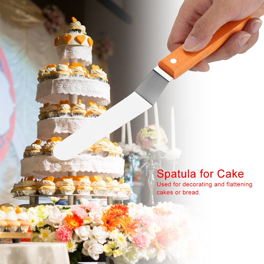 Eine Hand hält einen Spatel vor einer mehrstöckigen Torte und Blumen.