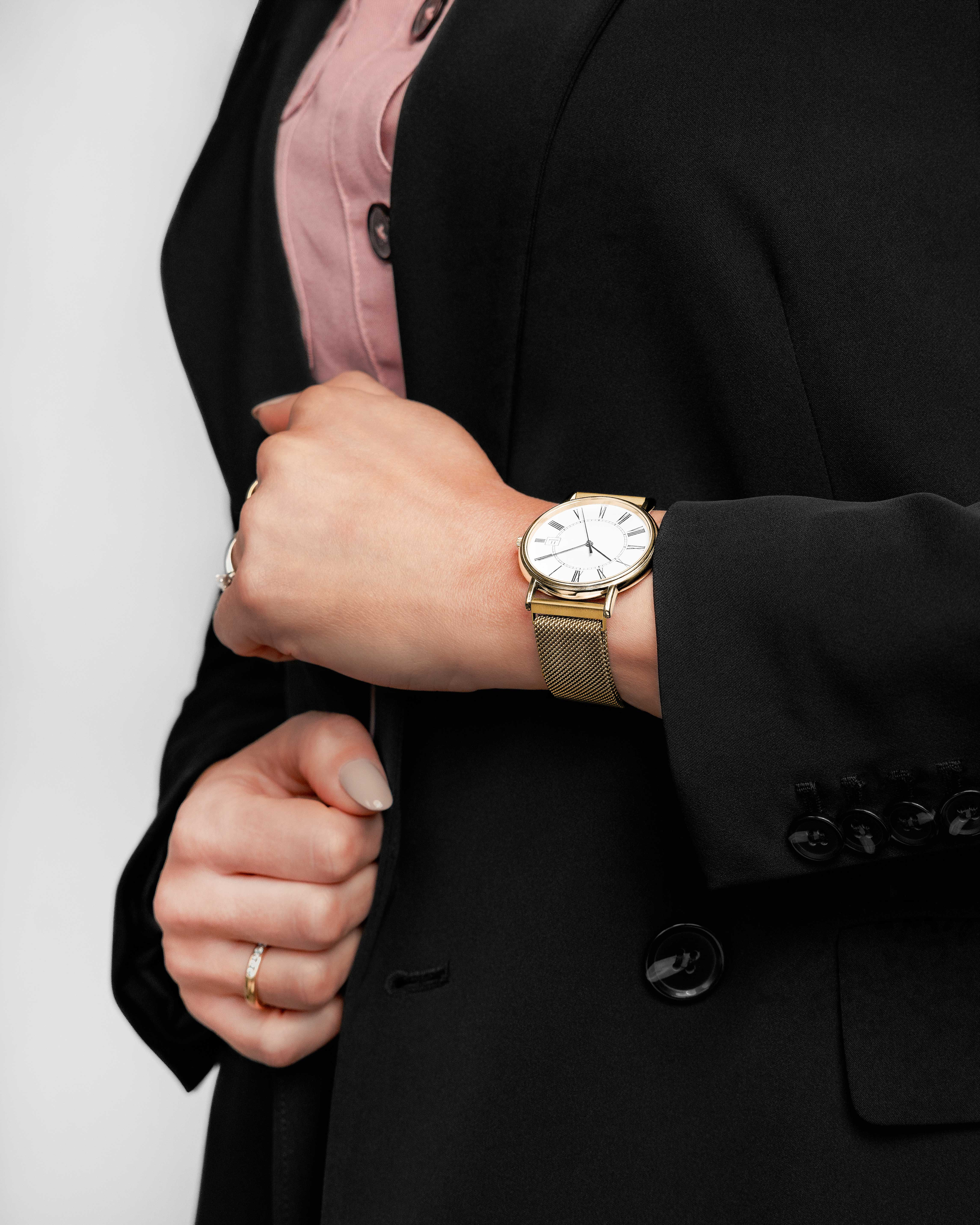 Person in schwarzem Blazer mit goldener Uhr. Rosa Hemd sichtbar. Hand mit Ringen. Neutraler Hintergrund.