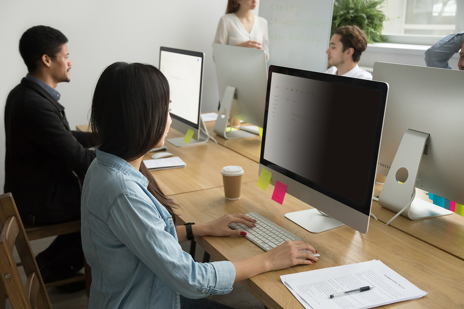 Scena biurowa. Ludzie przy biurkach z komputerami, klawiaturami i papierami. Jedna kobieta pisze na maszynie. Inni są w tle.