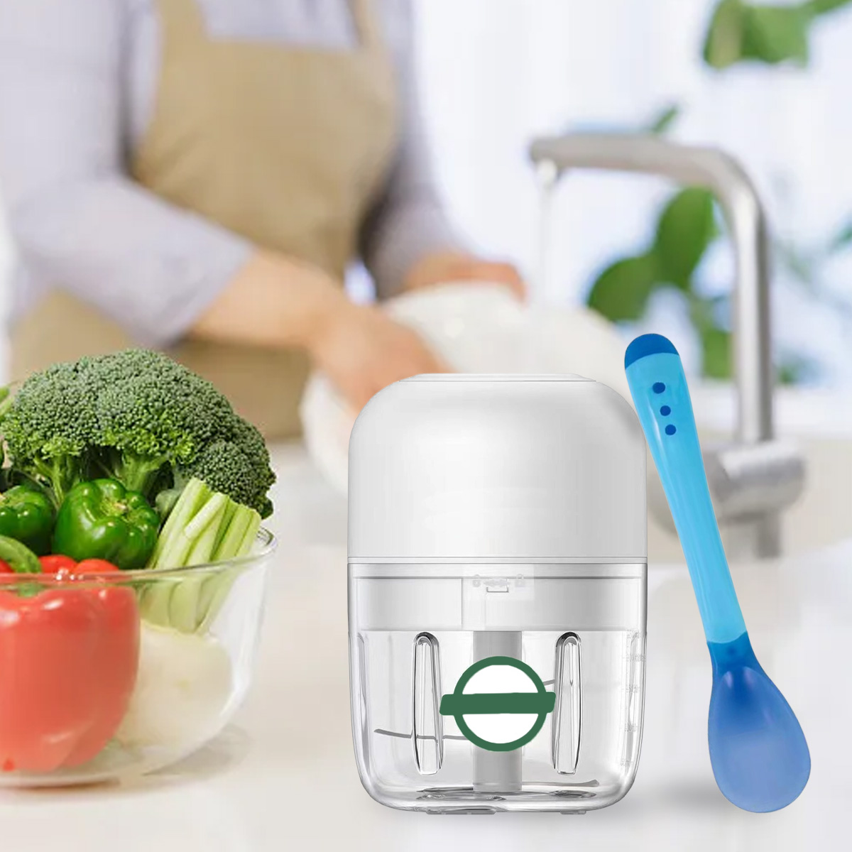 Ein weißer Food-Processor und blauer Löffel. Brokkoli, Paprika und rote Paprika in einer Glasschüssel. Eine Person wäscht im Hintergrund Geschirr.