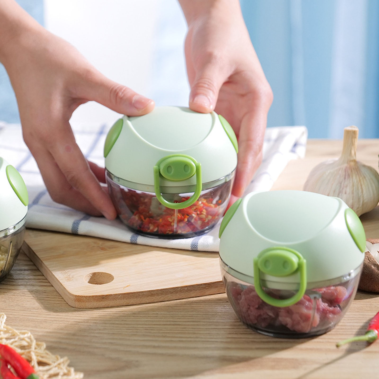 Hände halten grüne Zerkleinerer, einer mit gehackten roten Zutaten, einer mit Fleisch. Knoblauch und Chilischoten sichtbar.