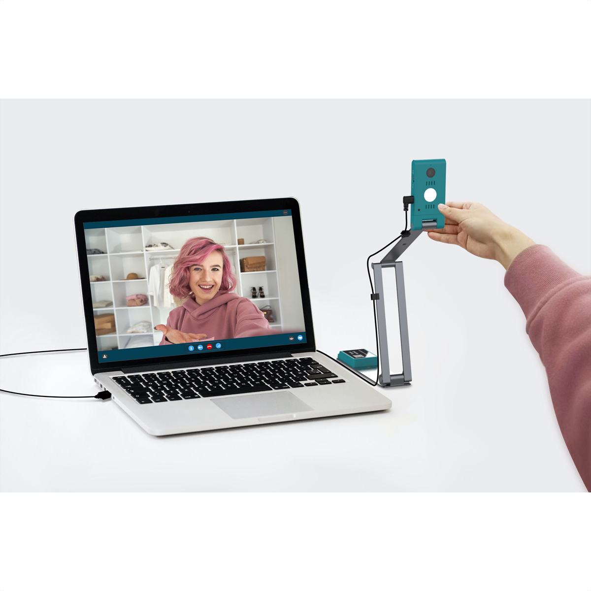 Ein Laptop zeigt eine Frau in einem Videoanruf. Eine Hand hält ein türkisfarbenes Gerät. Ein grauer Ständer und ein kleines Gerät sind ebenfalls vorhanden.