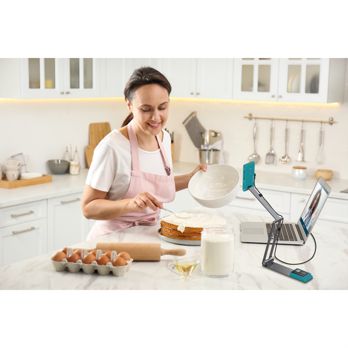Eine Frau in rosa Schürze, die einen Kuchen in einer Küche mit Eiern, Mehl und einem Laptop mit Videoanruf verziert.