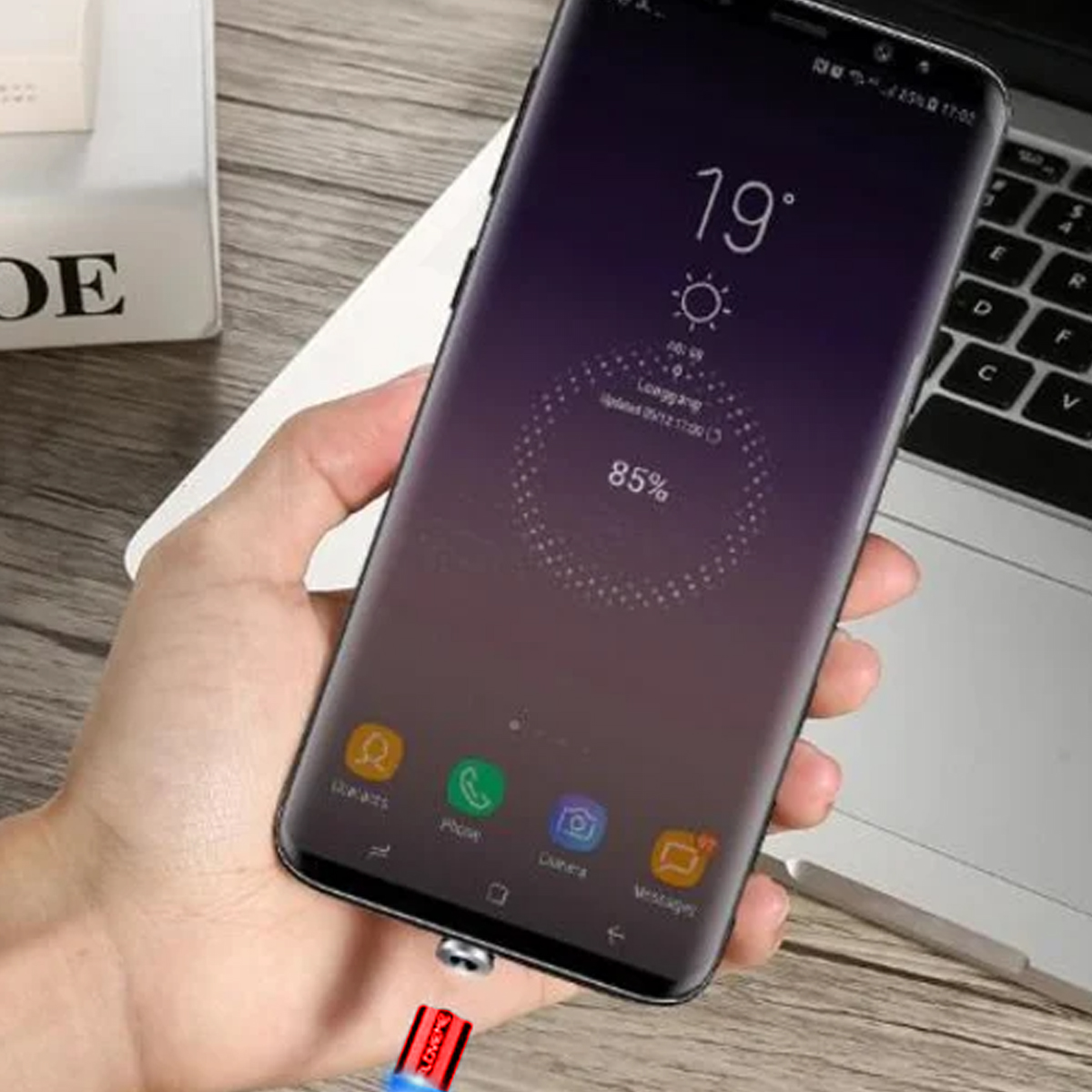 Hand hält Smartphone mit Ladekabel. Der Bildschirm zeigt 19 Grad, 85 % Akku und App-Symbole. Im Hintergrund ist ein Laptop.