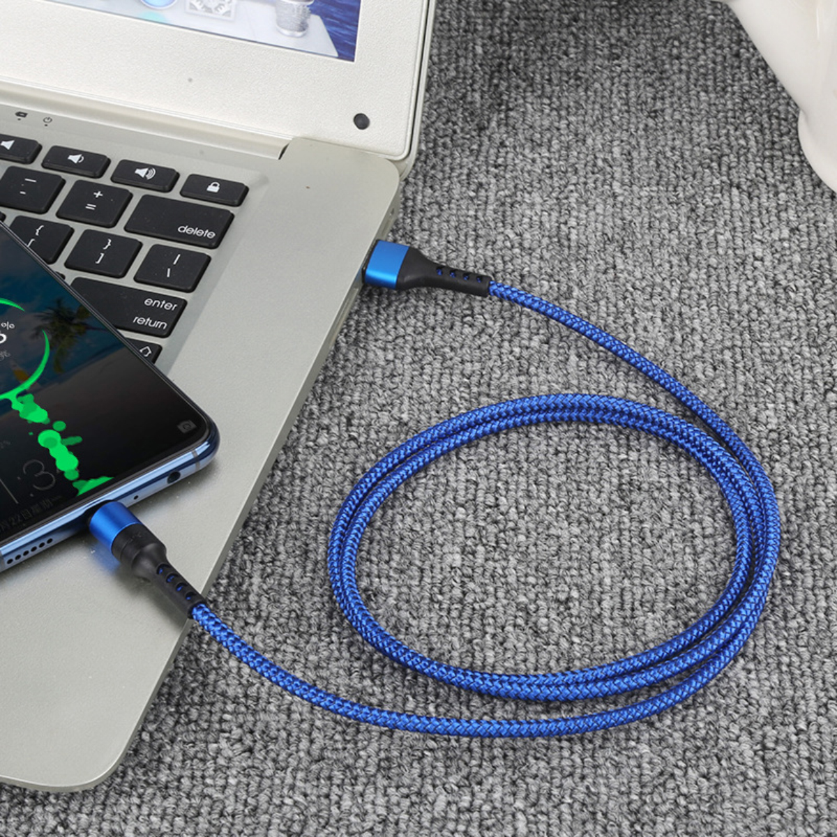 Ein blau geflochtenes USB-Kabel verbindet einen Laptop, ein Telefon. Das Telefon zeigt einen Ladebildschirm. Das Kabel liegt auf einem grauen Teppich.