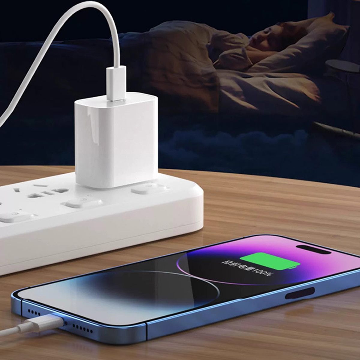 Ein Telefon, das auf einem Holztisch geladen wird. Ein weißes Ladegerät ist an eine Steckdosenleiste angeschlossen. Im Hintergrund schläft eine Person.