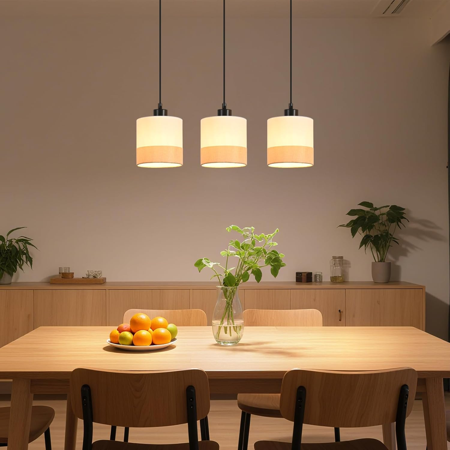 Sala da pranzo con tavolo in legno, sedie, fruttiera e vaso con piante. Tre lampade a sospensione sopra.
